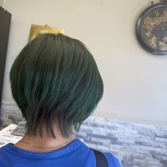 Woman with short, dark green hair, blue shirt, standing in a salon.