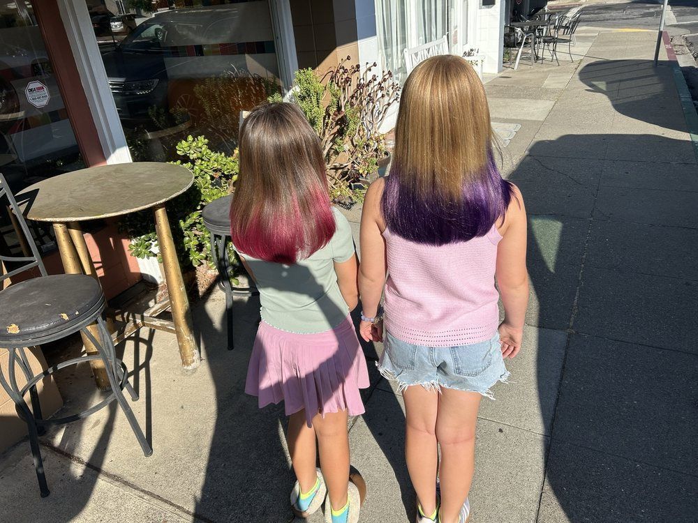 Young girl with pink and blue hair, smiling in a salon, wearing overalls and a white shirt.
