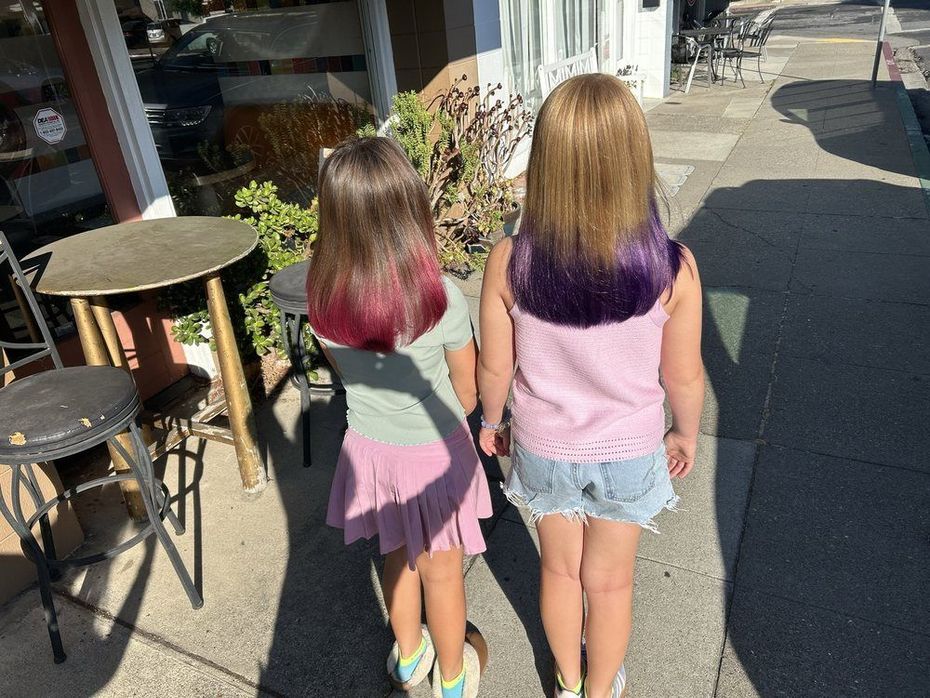 Young girl with pink and blue hair, smiling in a salon, wearing overalls and a white shirt.