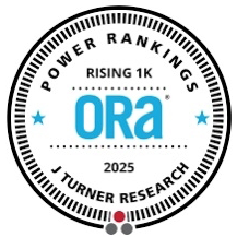 A small, brown, long-haired dog sits on a bed, looking towards the camera with a tilted head and an attentive expression.