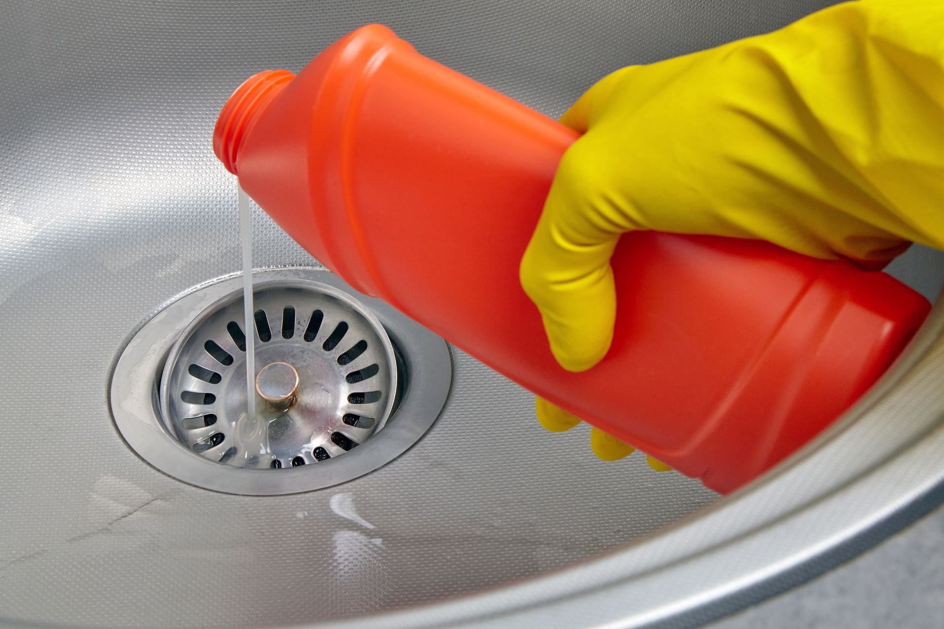 Hand in yellow glove pouring red liquid from bottle into kitchen sink drain.