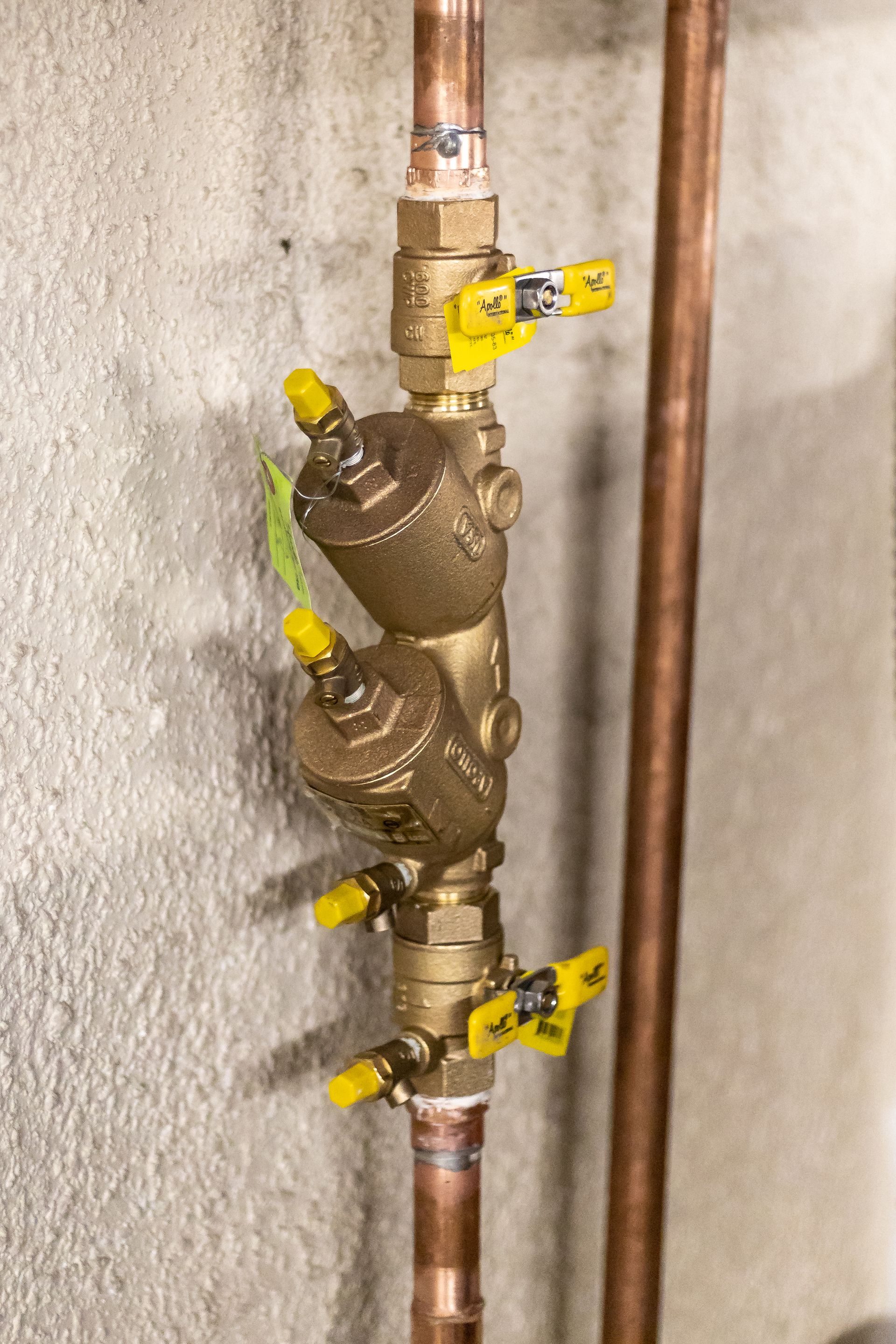 Copper pipes with brass backflow preventer and four yellow-handled ball valves on a gray wall.