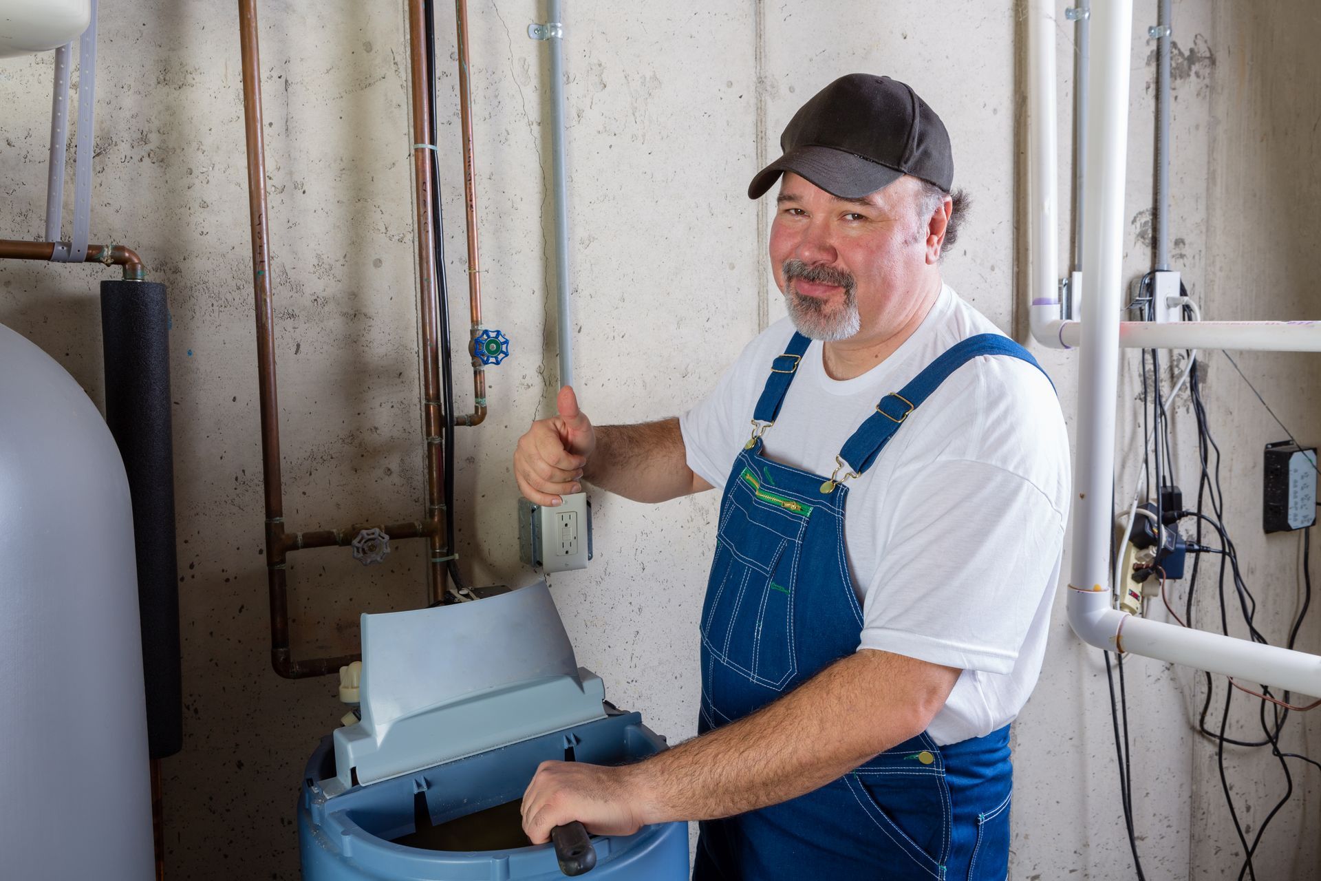 Man in overalls giving a thumbs-up next to a water softener in a utility room.