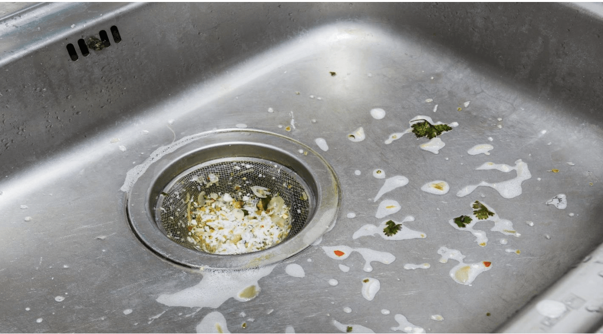 A dirty stainless steel kitchen sink with food stuck in the drain.