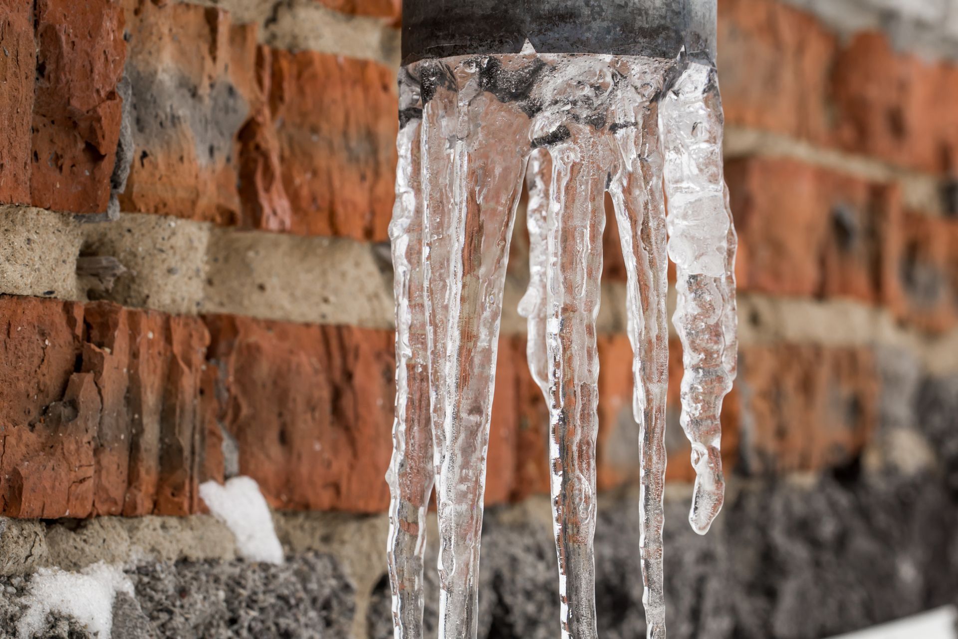 Icicles are hanging from a pipe in front of a brick wall.