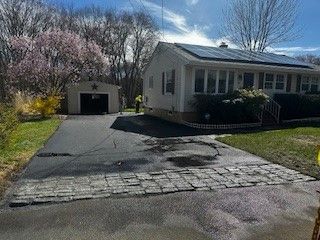 Asphalt driveway with damaged brick patch, leading to a ranch-style house with solar panels. Garage to the left.