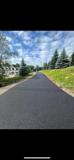 A freshly paved road stretches into the distance, flanked by green grass, pine trees, and a cloudy sky.