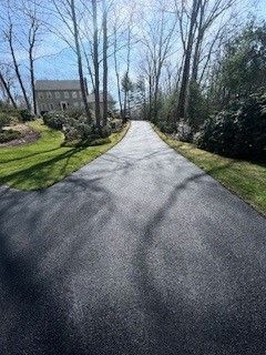 Long, dark asphalt driveway leading toward a house through a tree-lined area on a sunny day.