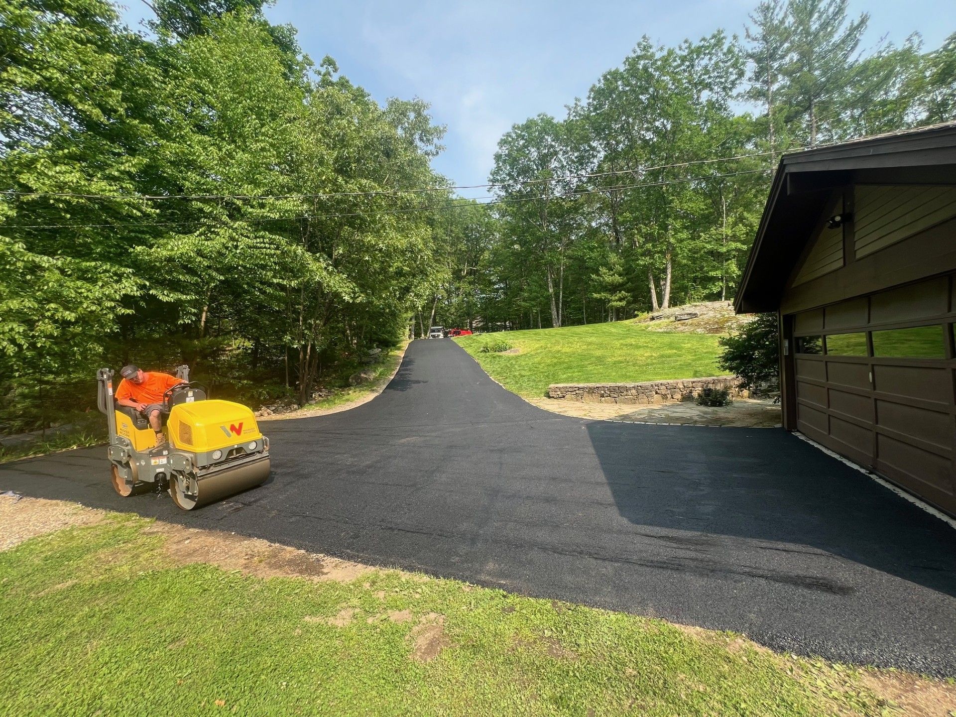 Asphalt driveway being rolled by a compact vibratory roller next to a garage, trees in the background.
