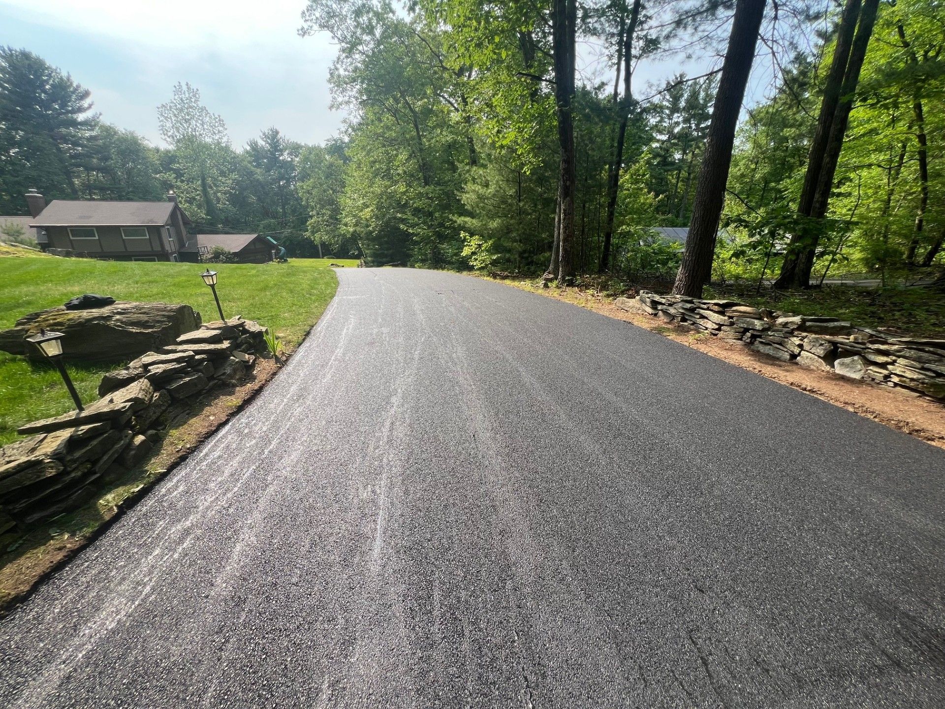 Newly paved asphalt driveway leading toward a house amidst trees on a sunny day.