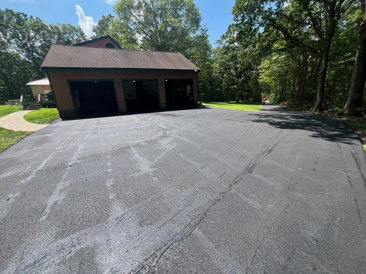 Black asphalt driveway leading to a three-bay garage, surrounded by trees under a blue sky.