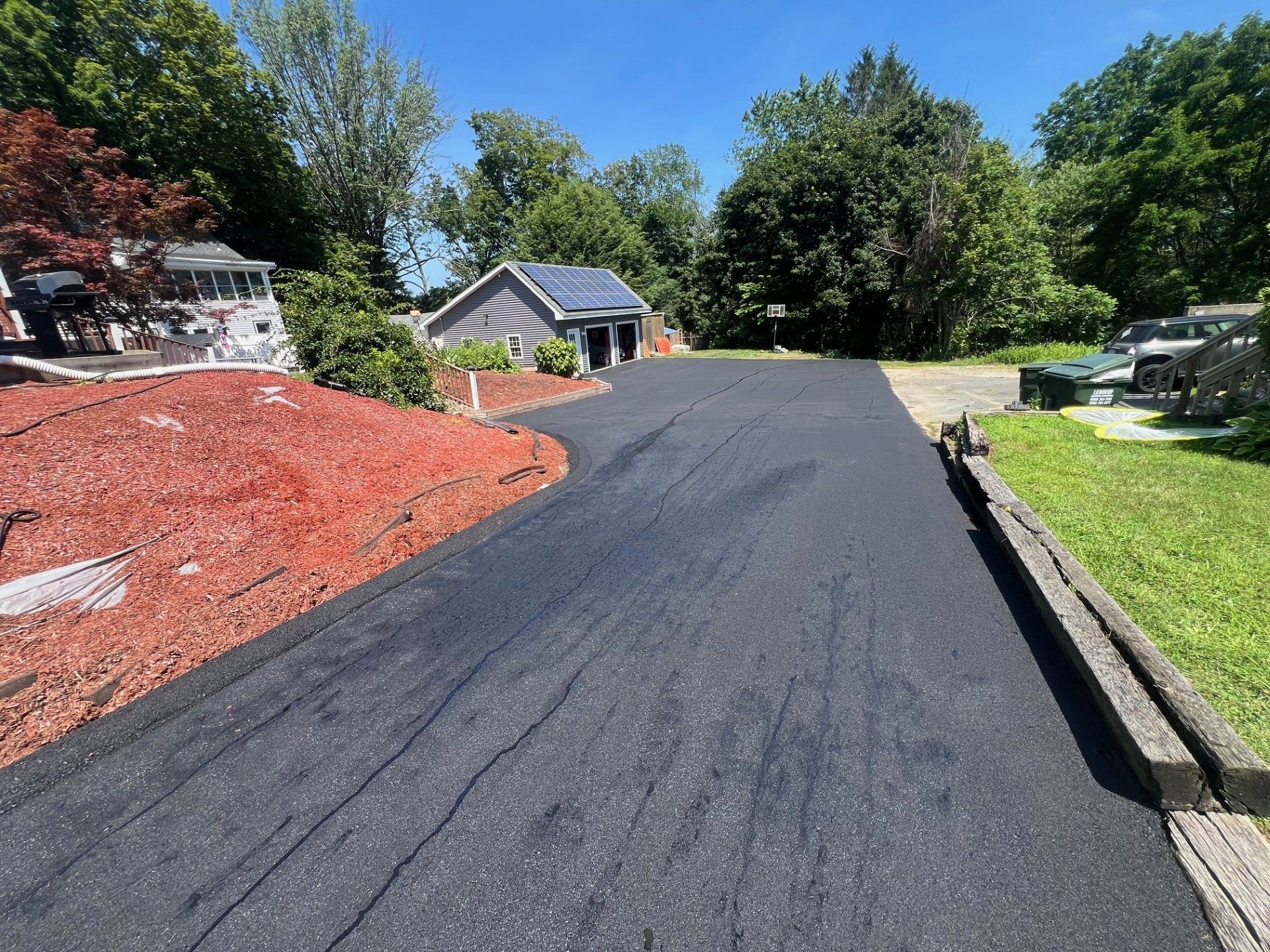Black asphalt driveway curves toward a garage under a blue sky, flanked by red mulch and grass.
