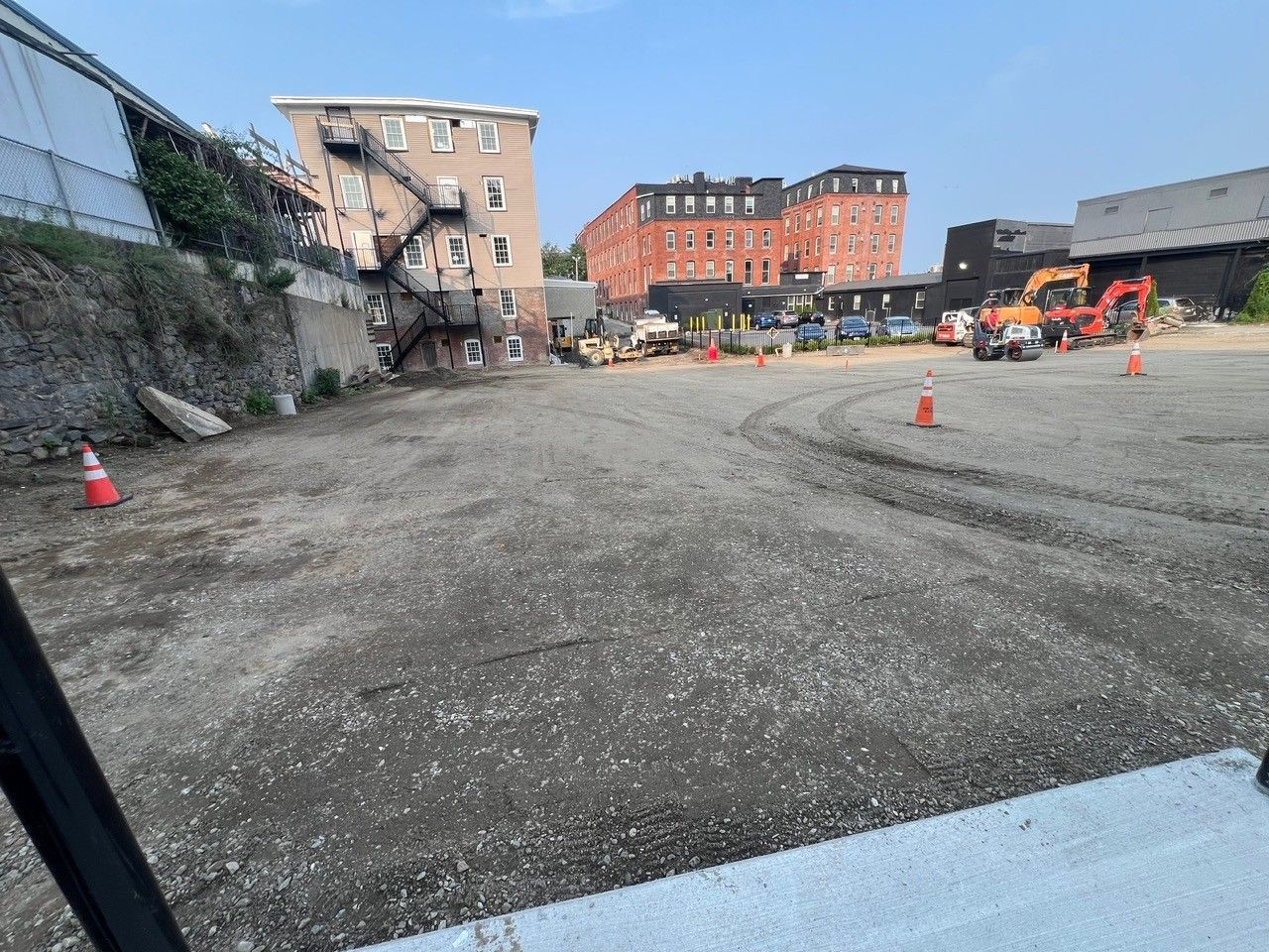 A gravel lot with construction equipment and buildings in the background under a blue sky.