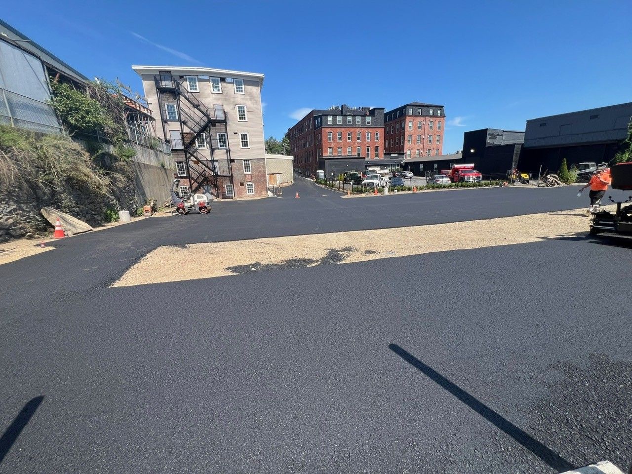Newly paved parking lot with buildings in the background and gravel border on a sunny day.
