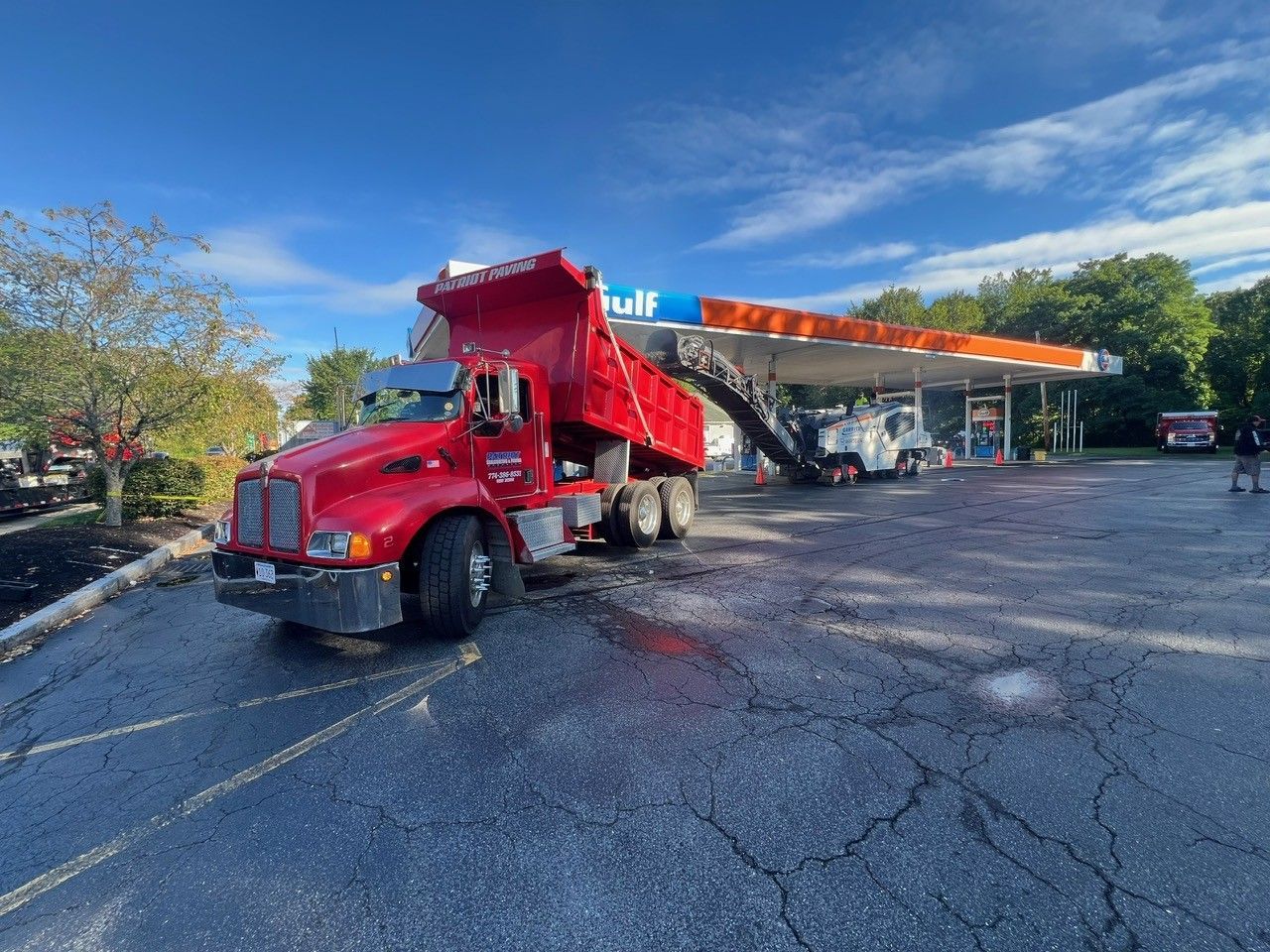 Red dump truck unloading asphalt at a Gulf gas station under a blue sky.