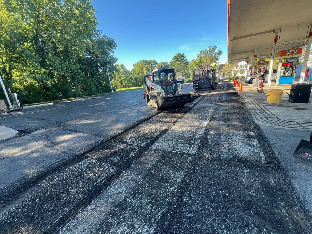 Asphalt paving project at a gas station.  Skid steer loaders removing old pavement under a bright blue sky.