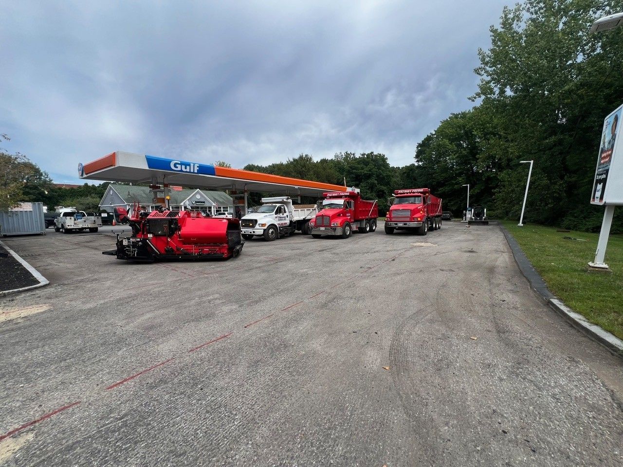 Construction vehicles at a gas station, including a red asphalt grinder, trucks, and a white pickup. Overcast sky.