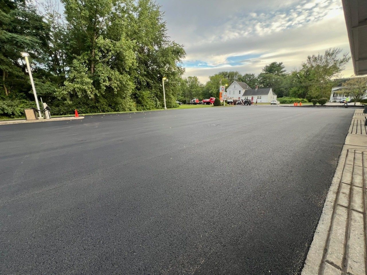 Freshly paved black asphalt parking lot with trees and a building in the background.
