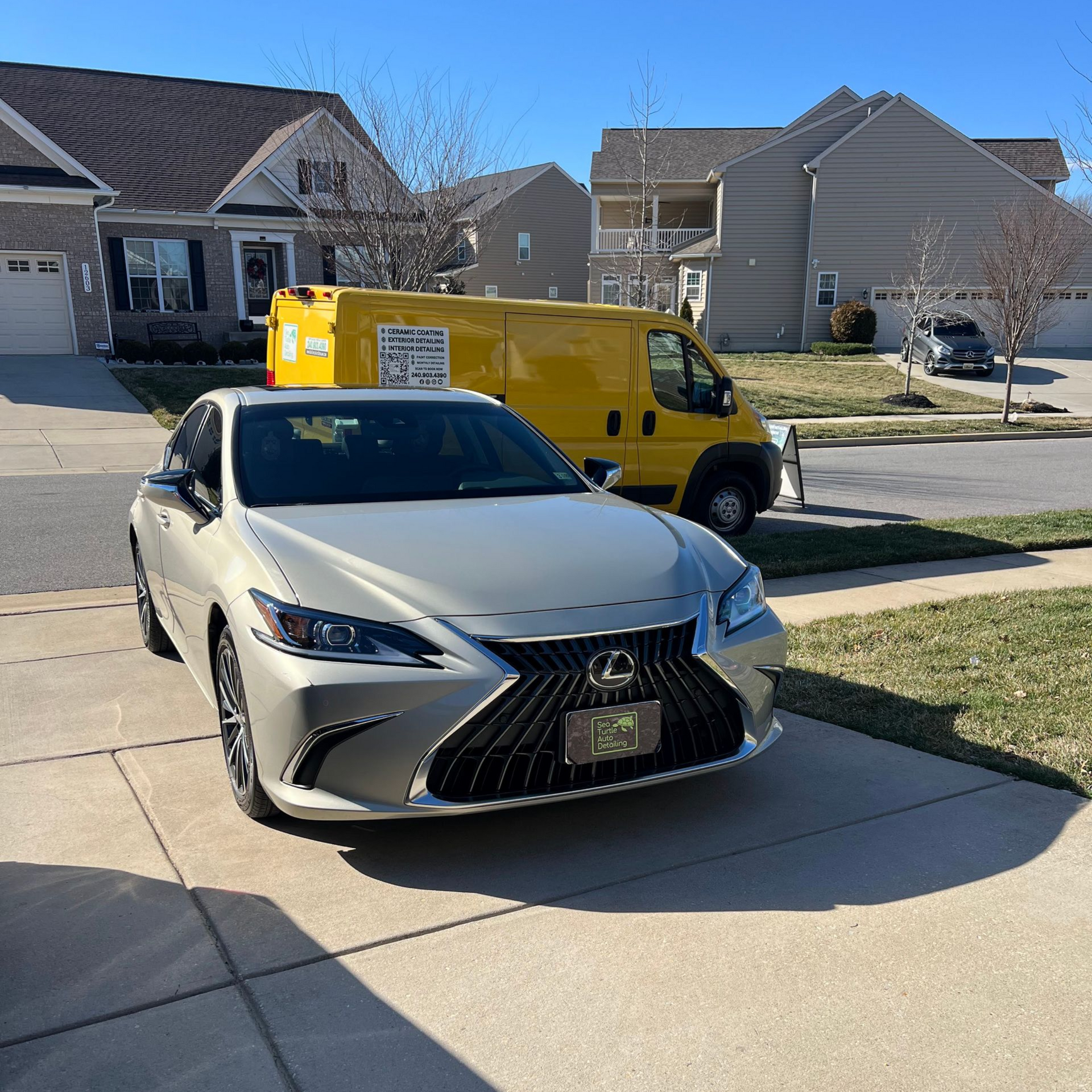 A silver Lexus parked in a residential driveway with a yellow utility van parked on the street behind it.
