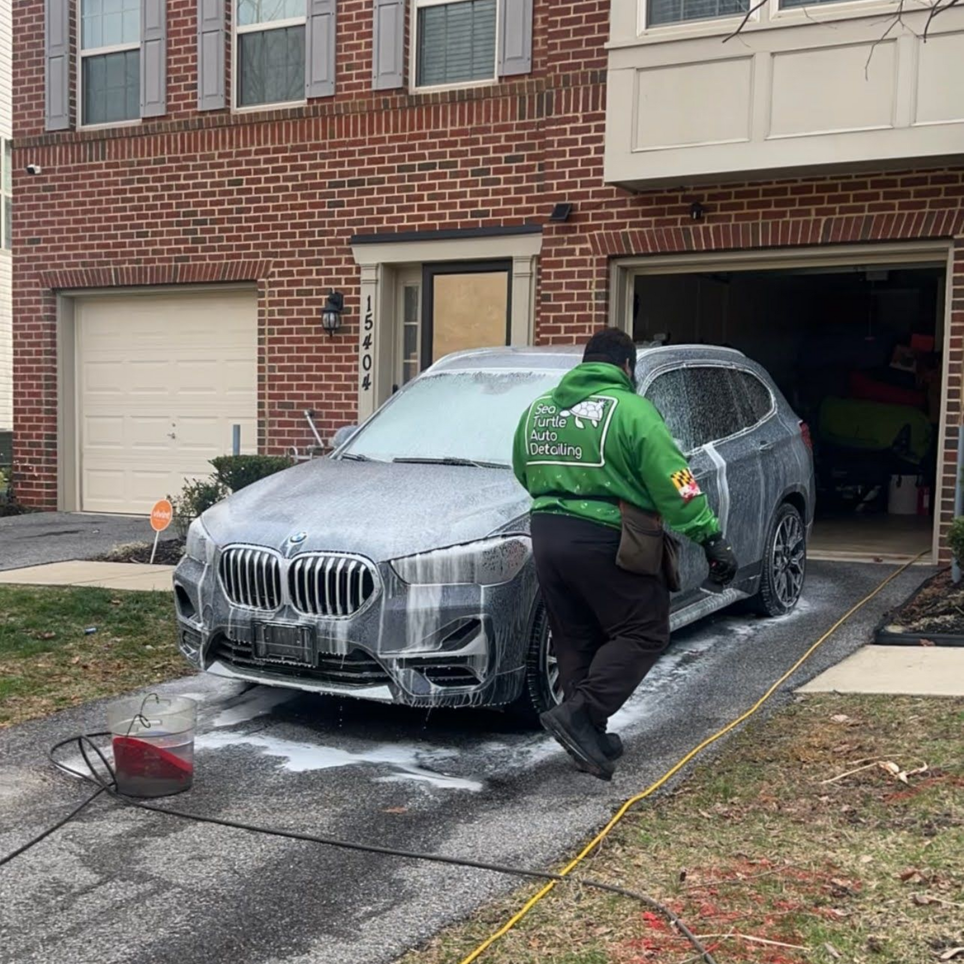 A person in a bright green hoodie uses soapy foam to wash a dark BMW SUV parked in a residential driveway.