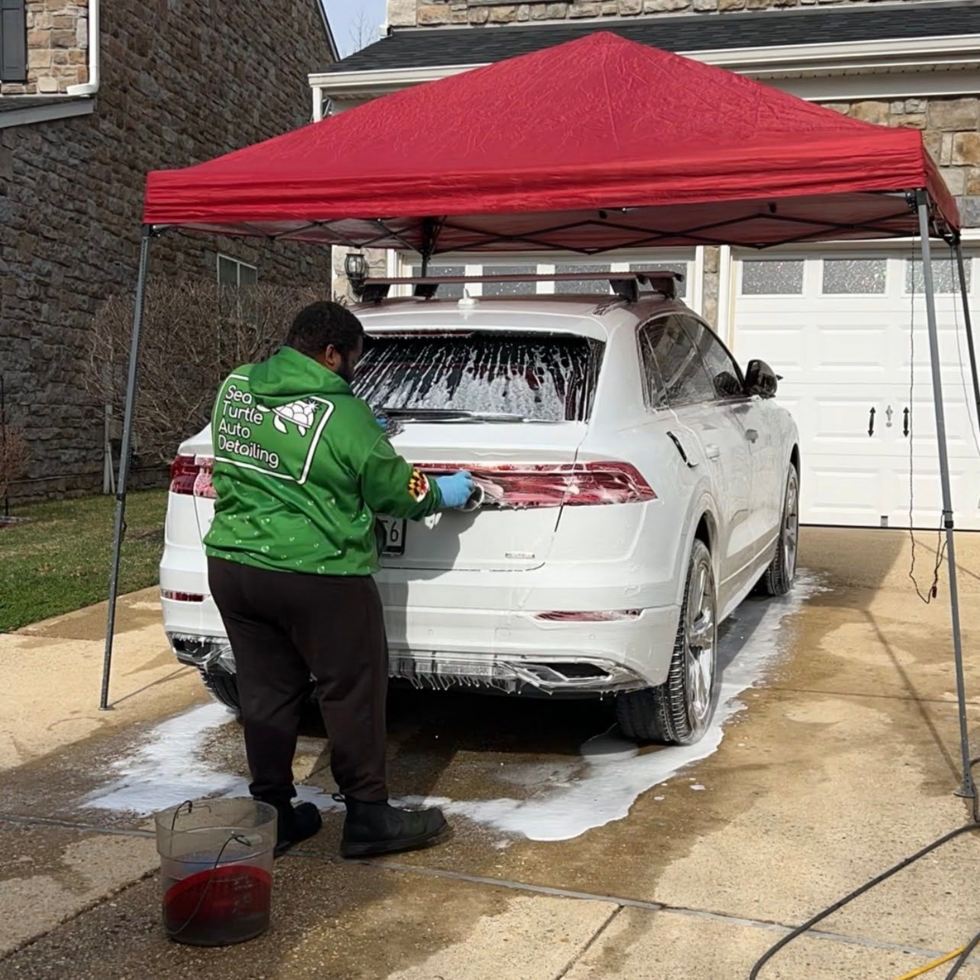 A person in a green hoodie washes a white car under a red canopy in a driveway.