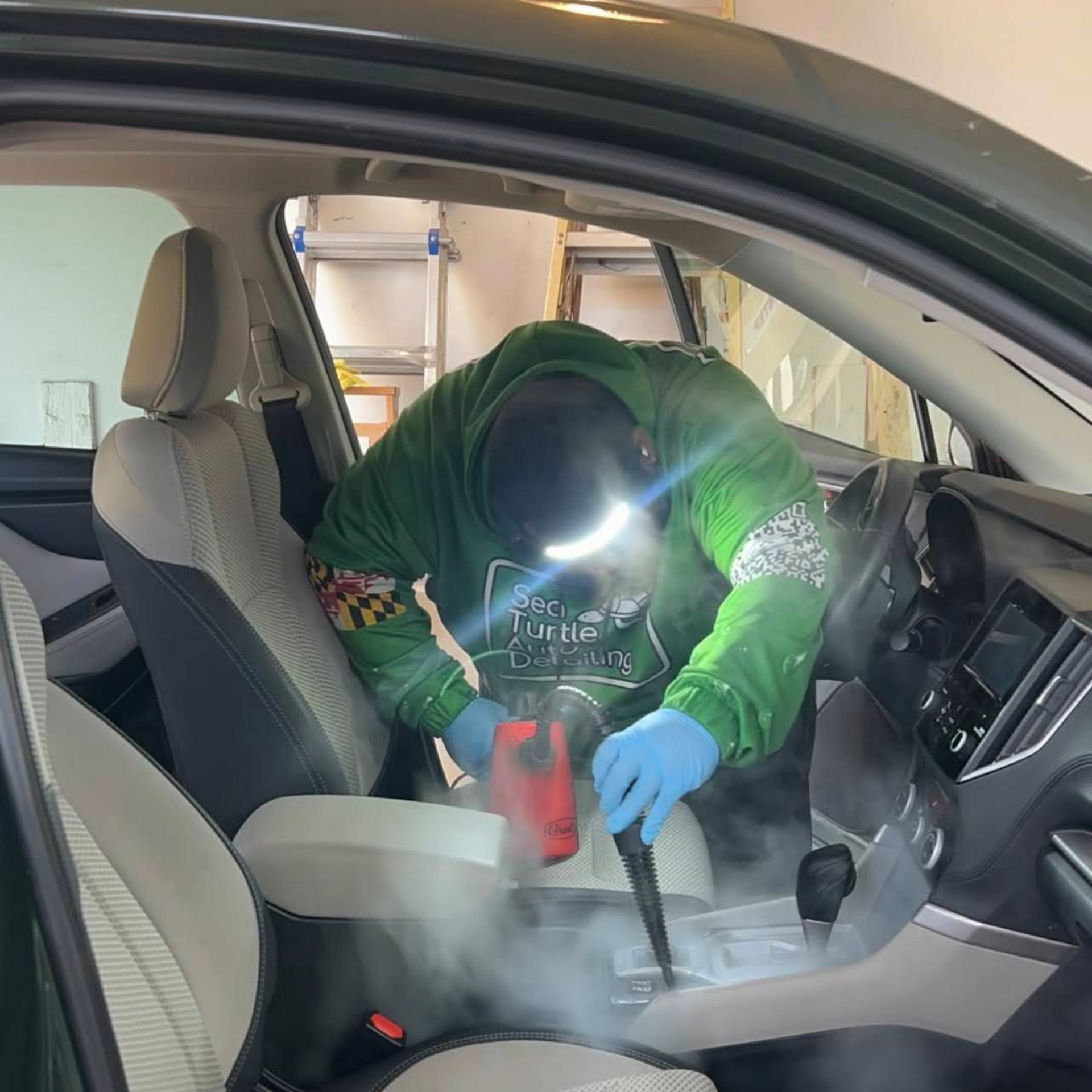 A person in a green hoodie wearing blue gloves steam cleans the center console of a car interior.
