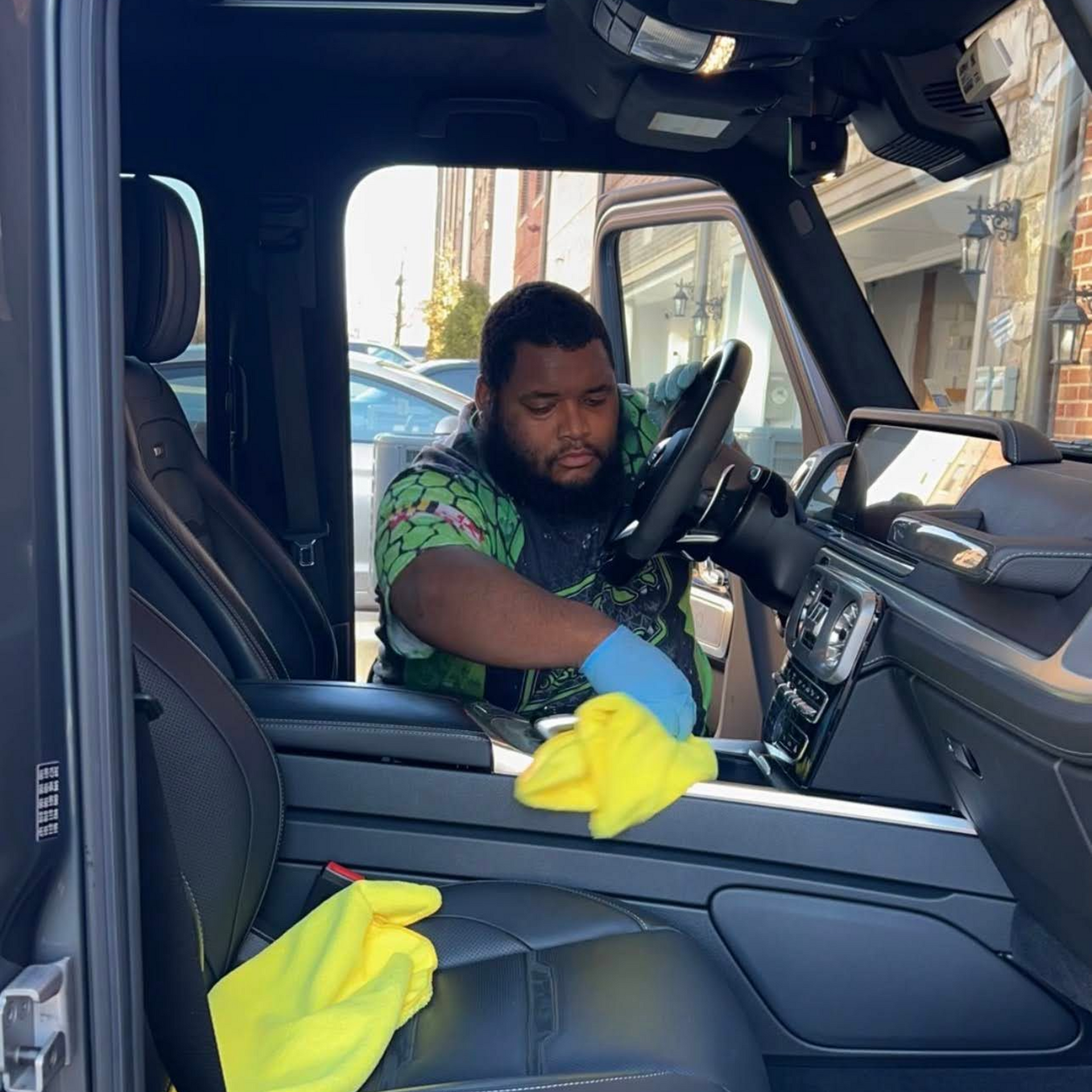 A person wearing a patterned shirt and blue gloves cleans the interior of a vehicle with a yellow microfiber cloth.