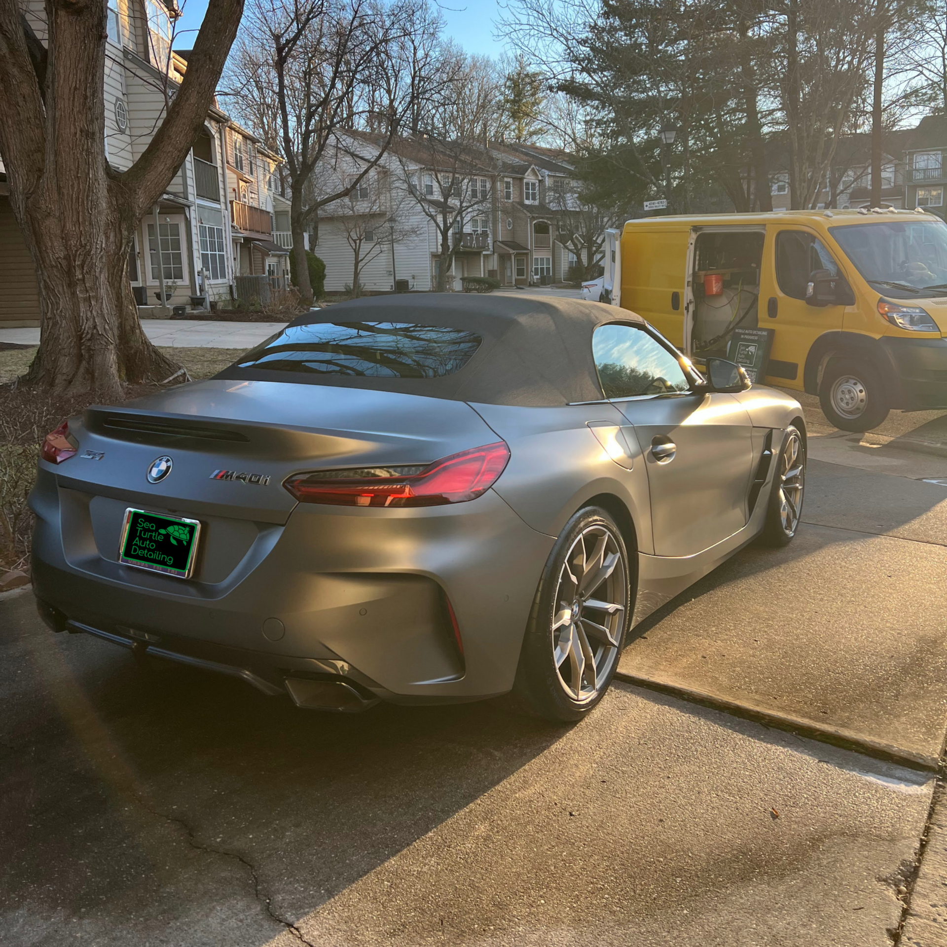 A gray BMW Z4 convertible parked on a suburban street next to a yellow utility van on a sunny day.