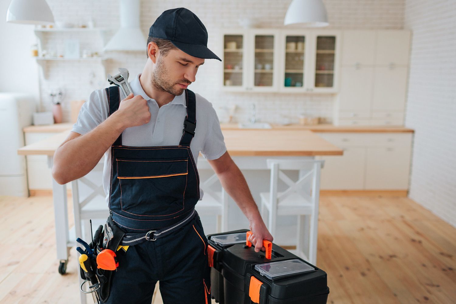 Plumber in overalls and cap holding a wrench, standing in a kitchen with a toolbox.