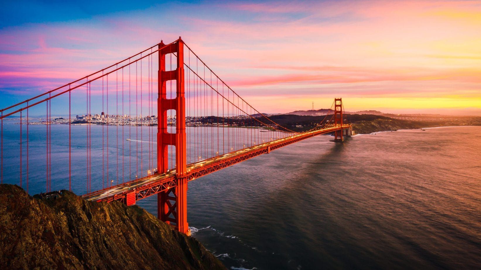 Golden Gate Bridge at sunset, orange-red against colorful sky and water.