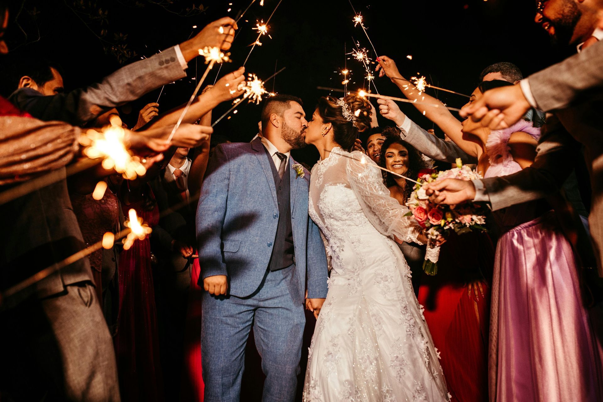Newly married couple kissing, surrounded by wedding guests holding sparklers, night.