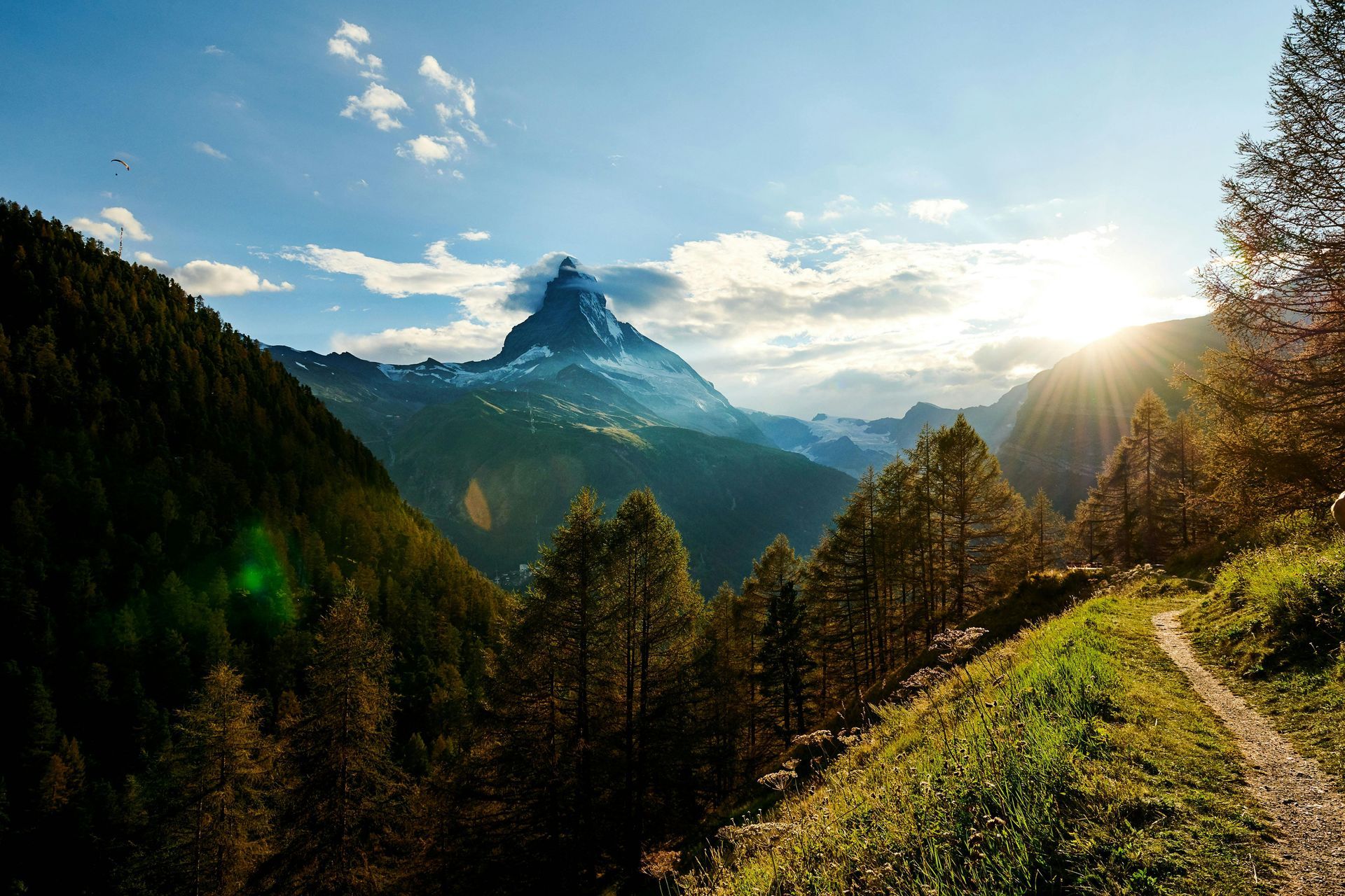 A trail winds through a valley towards the Matterhorn mountain, bathed in sunlight.