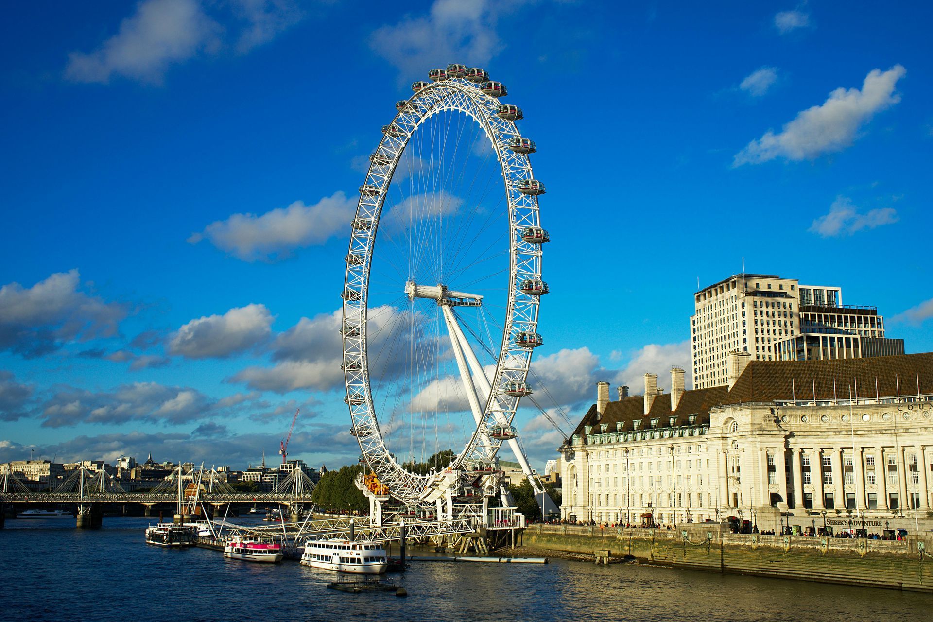 London Eye ferris wheel, Thames River, blue sky with clouds, buildings, boats.