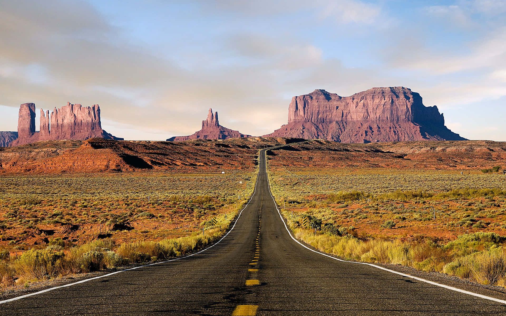 Road stretches towards red rock formations in desert landscape, under blue and cloudy sky.