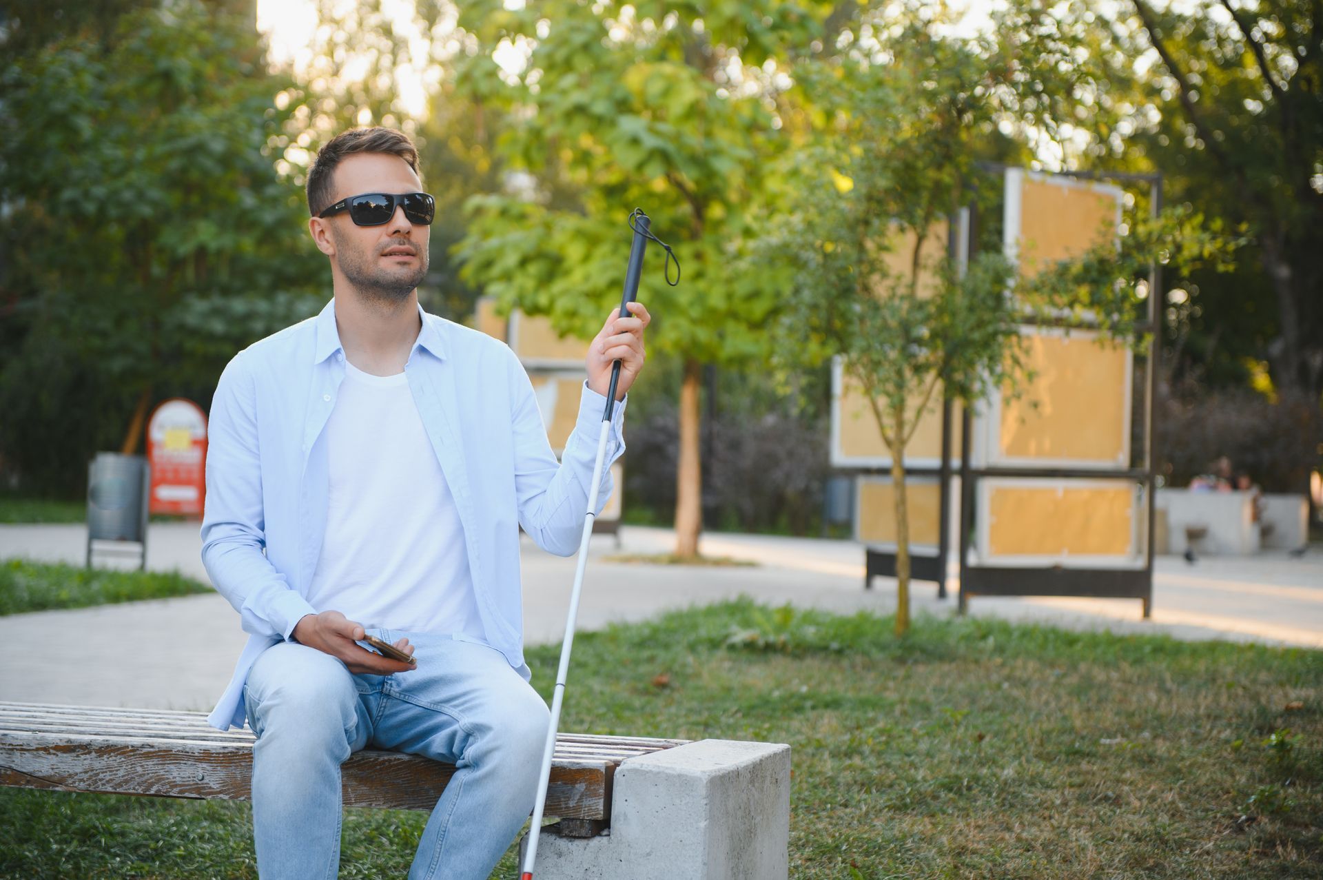 A blind man sitting on a bench in the park with a cane