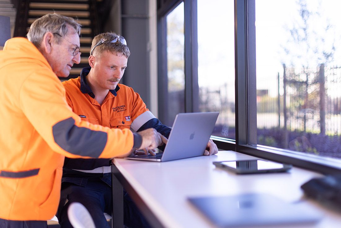 Two Men Are Sitting at a Table Looking at a Laptop Computer — Macquarie Electric in Brisbane, QLD