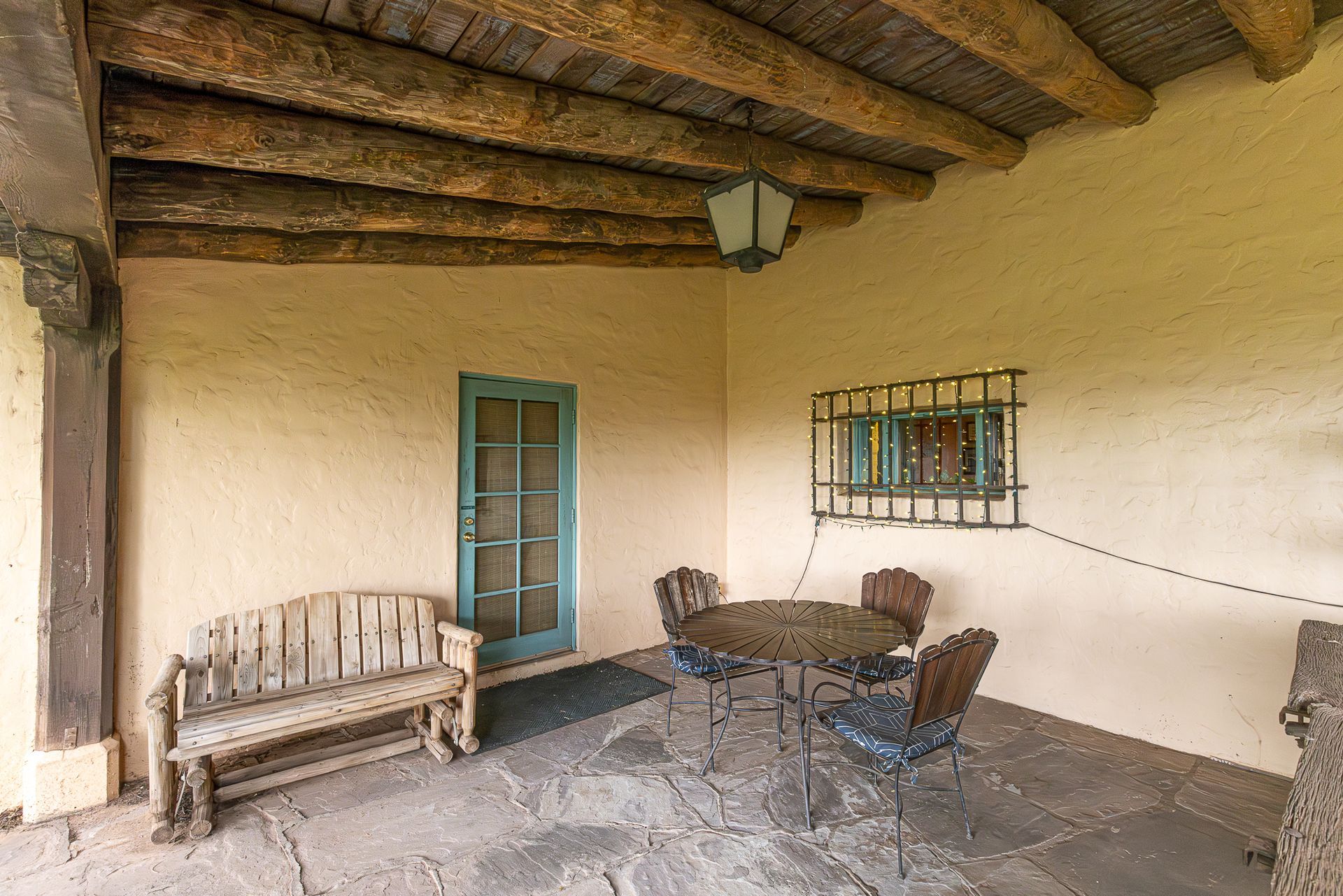 Covered patio with wooden bench, wrought-iron table and chairs. Turquoise door, window with grill, and wood-beam ceiling.