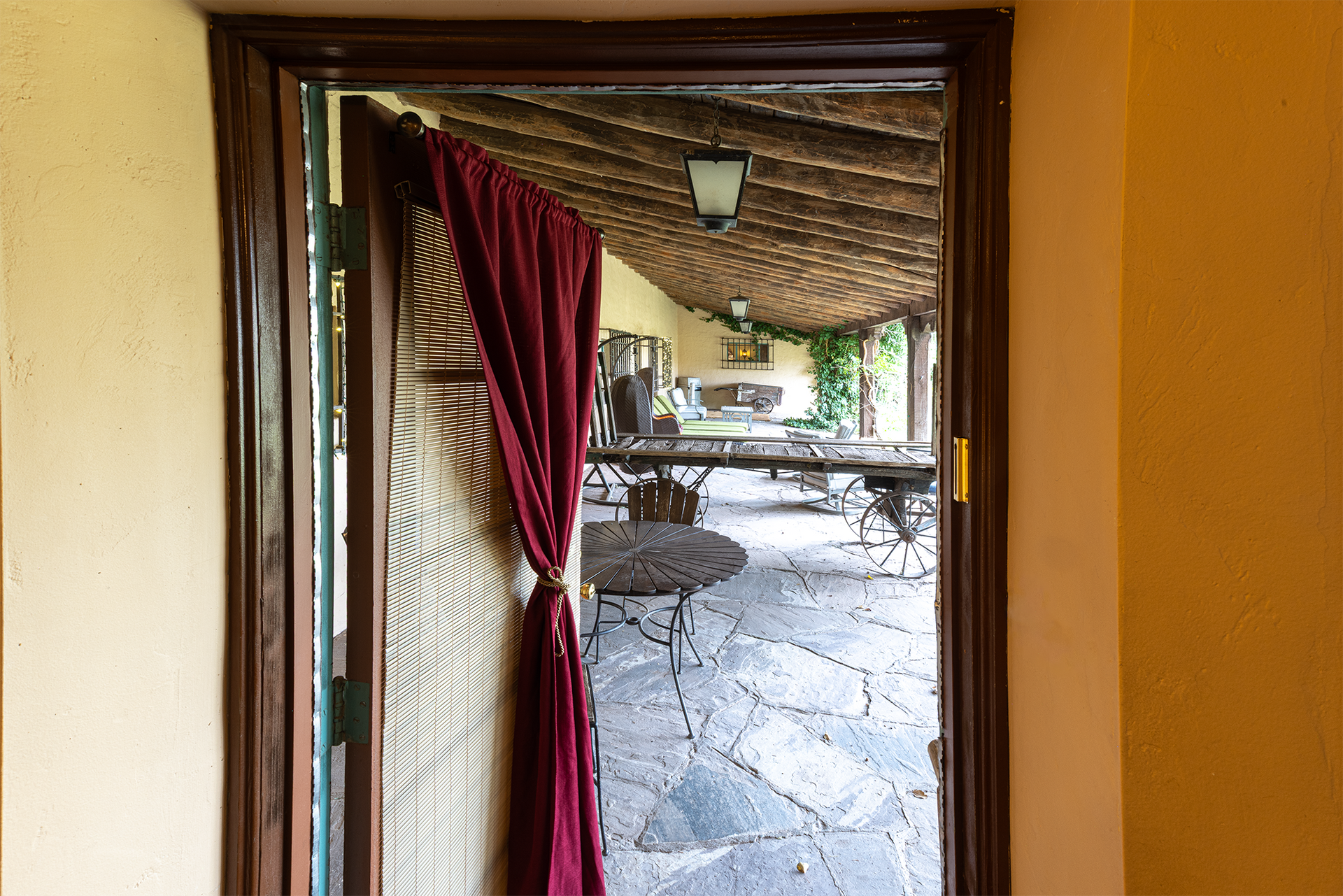 Doorway with a red curtain partially blocking the view to an outdoor patio with a stone floor and wooden roof.