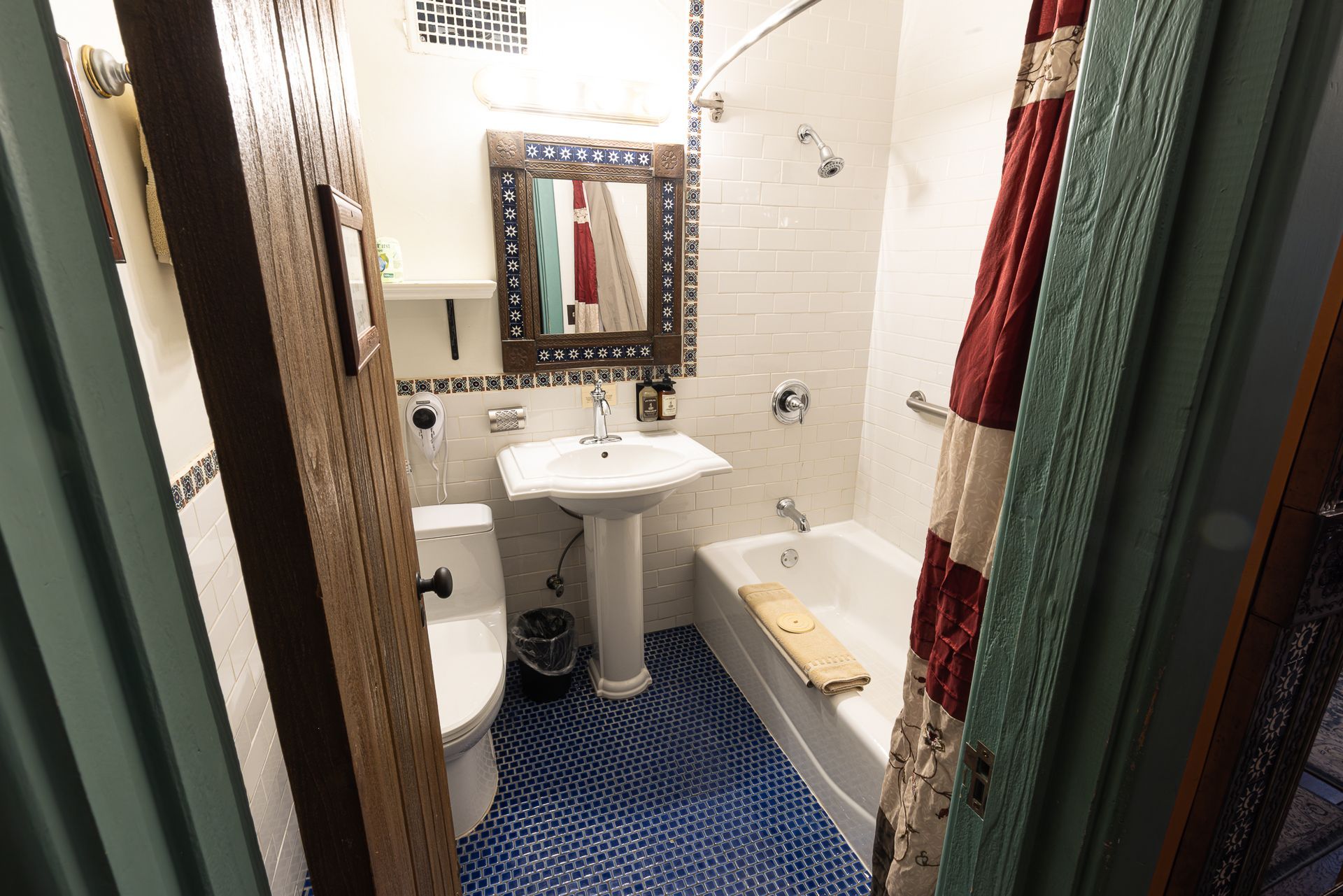 Small bathroom with a white toilet, sink, and bathtub. Blue tiled floor, patterned red shower curtain, and brown door frame.