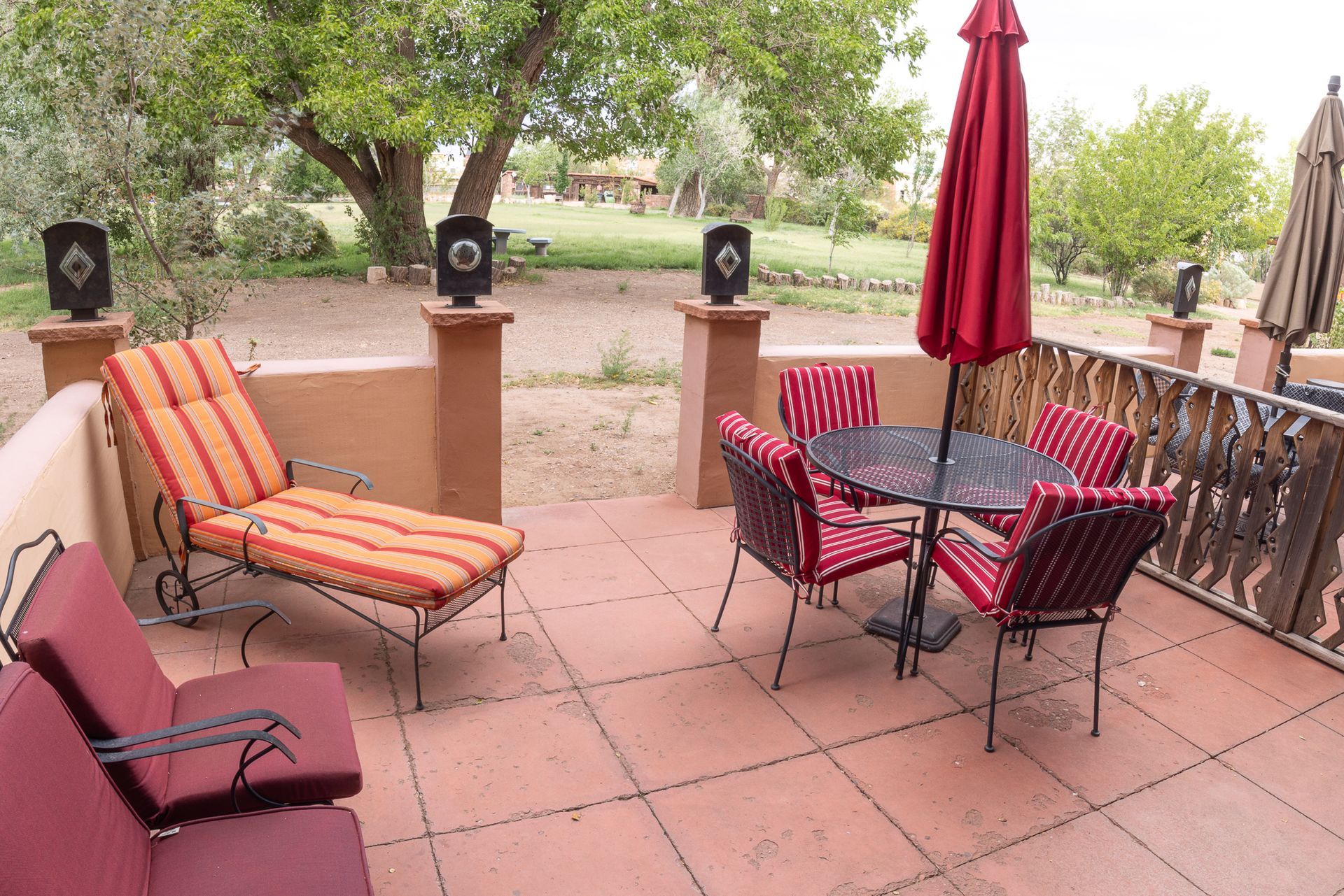 Patio with red chairs, table, umbrella. Lounge chair, red-tile floor, and brown wall in outdoor setting.
