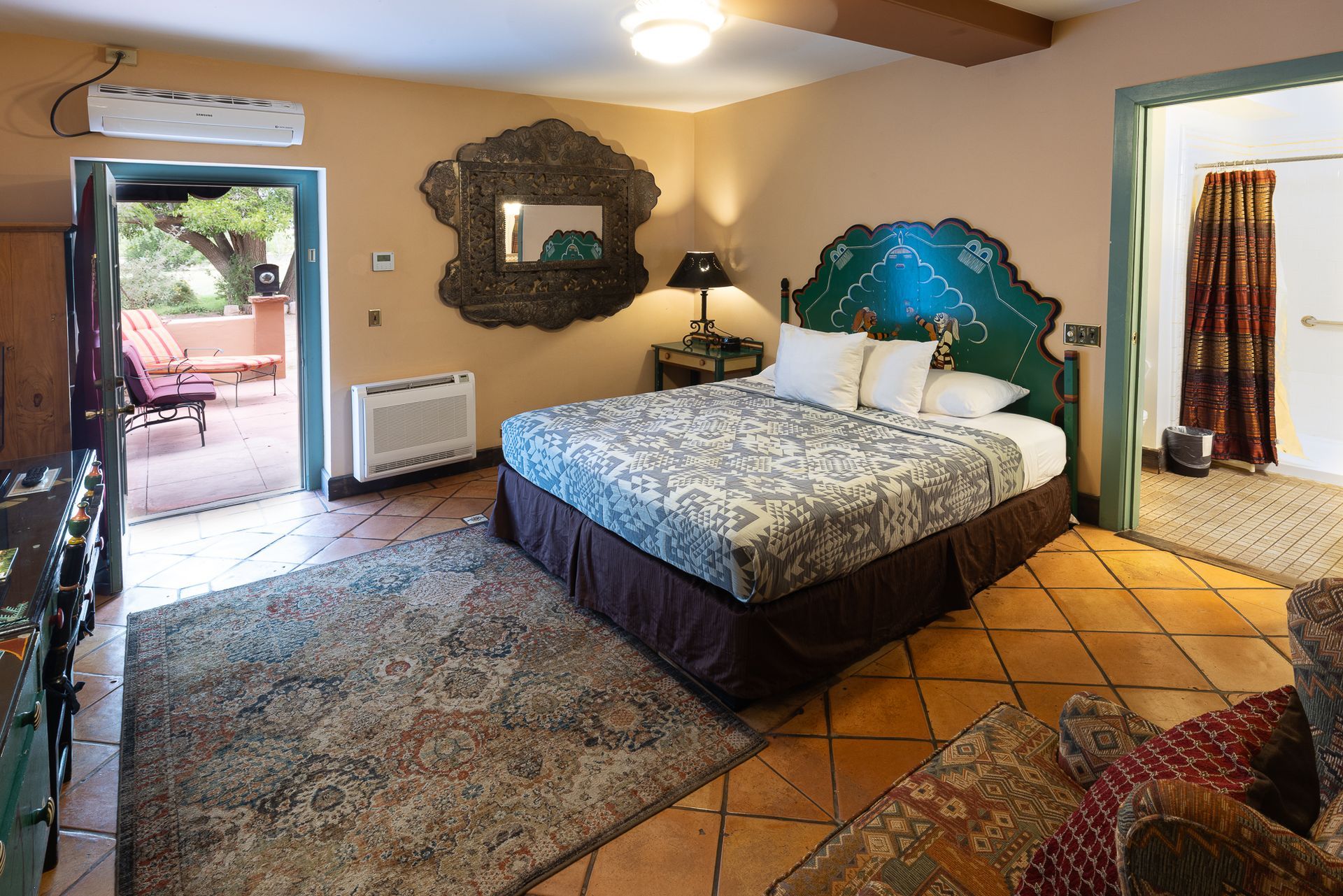 Bedroom with a bed, outdoor patio access, and decorative mirror. Beige walls, terra cotta floors, and colorful decor.
