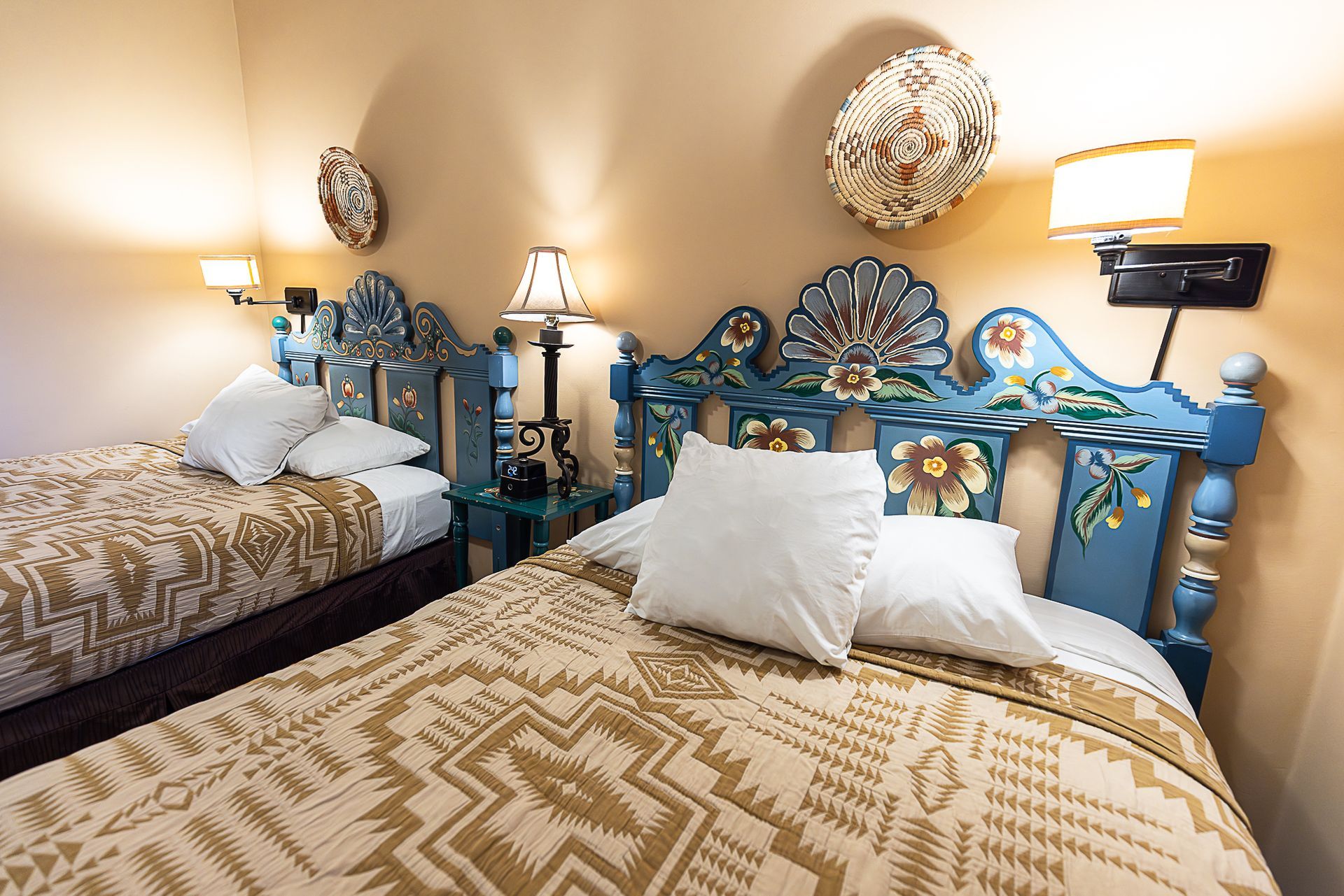 Two beds with blue, decorative headboards. Beige patterned blankets, white pillows, and lamps.