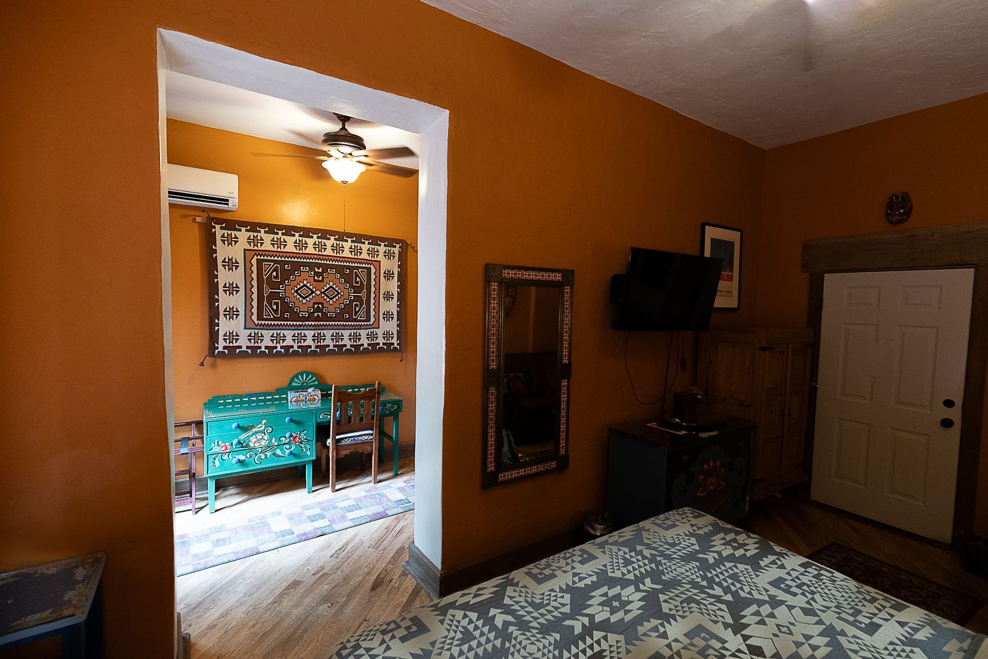 Room with orange walls, doorway to another room with colorful furniture, rug on the floor, and a TV on the wall.