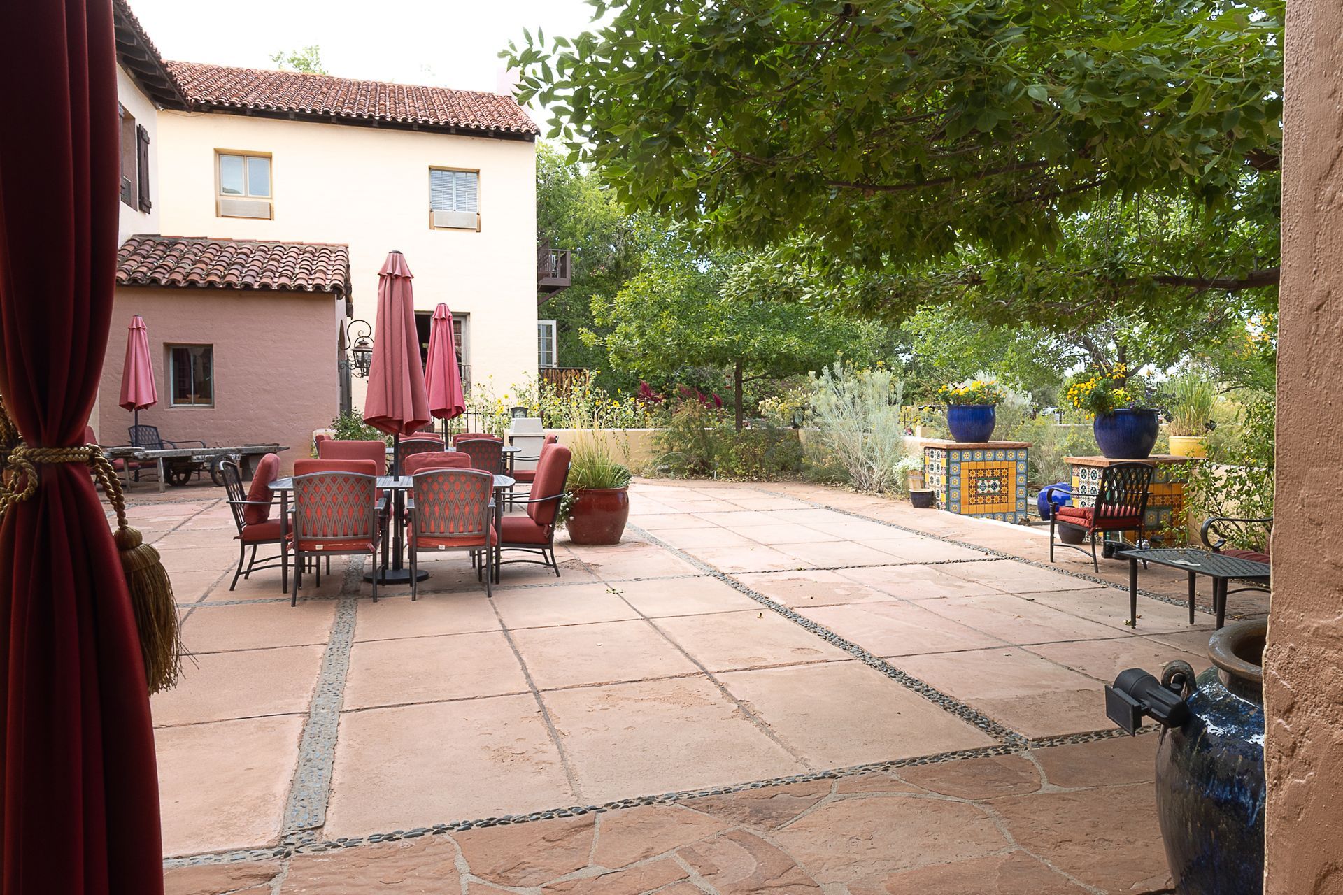Courtyard with patio furniture, red umbrellas, and a building with a red-tiled roof, plants, and trees.