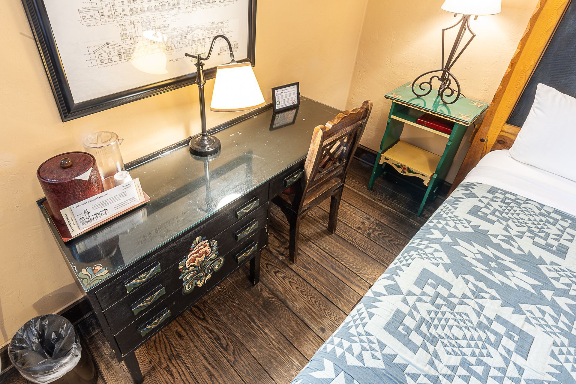 Desk with chair, lamp, and amenities in a hotel room with wood floors and a bed.