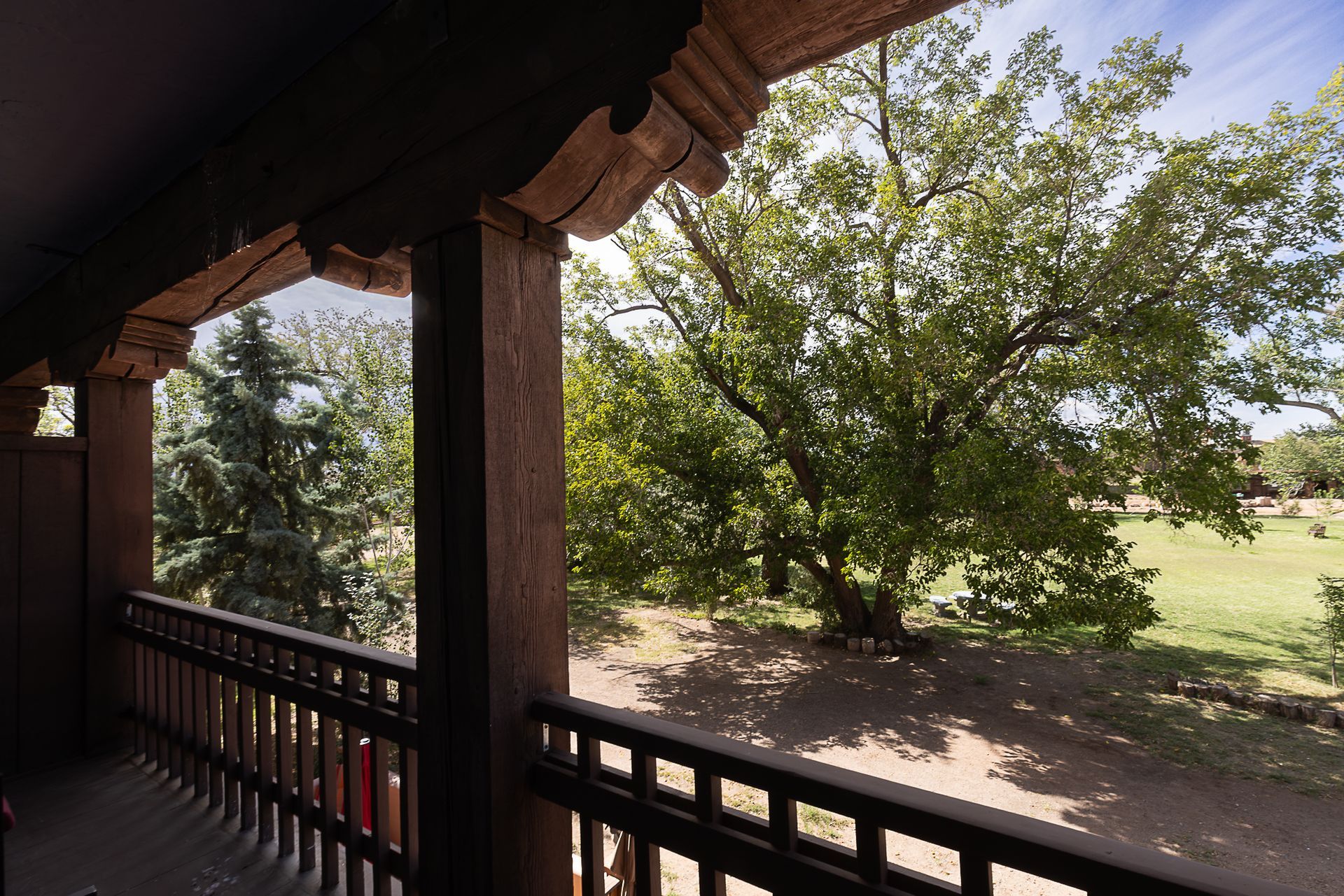 Balcony with carved wood details overlooking a green lawn and trees under a blue sky.