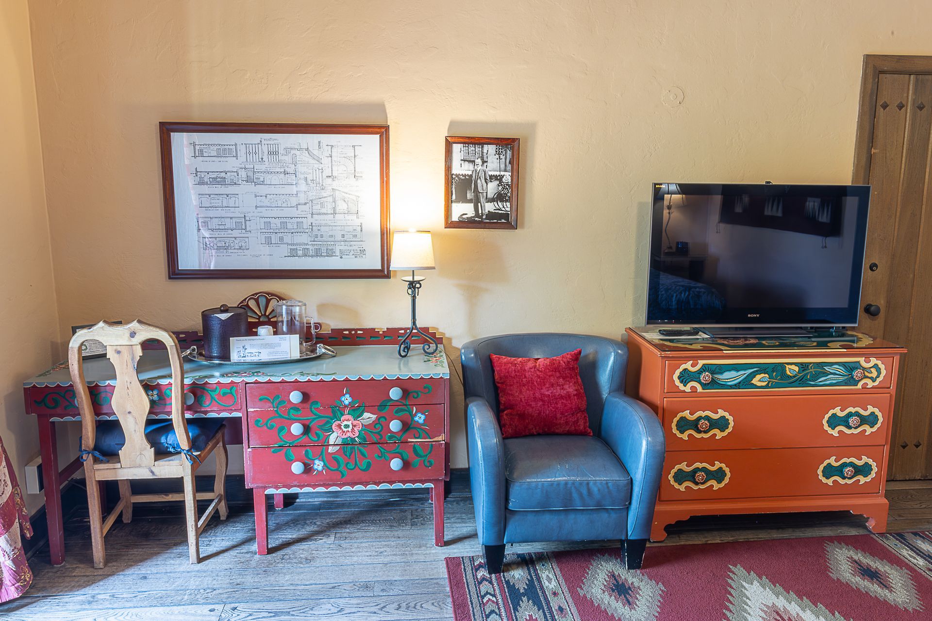 Room with desk, chair, TV on painted dresser, blue chair with red pillow, framed art.
