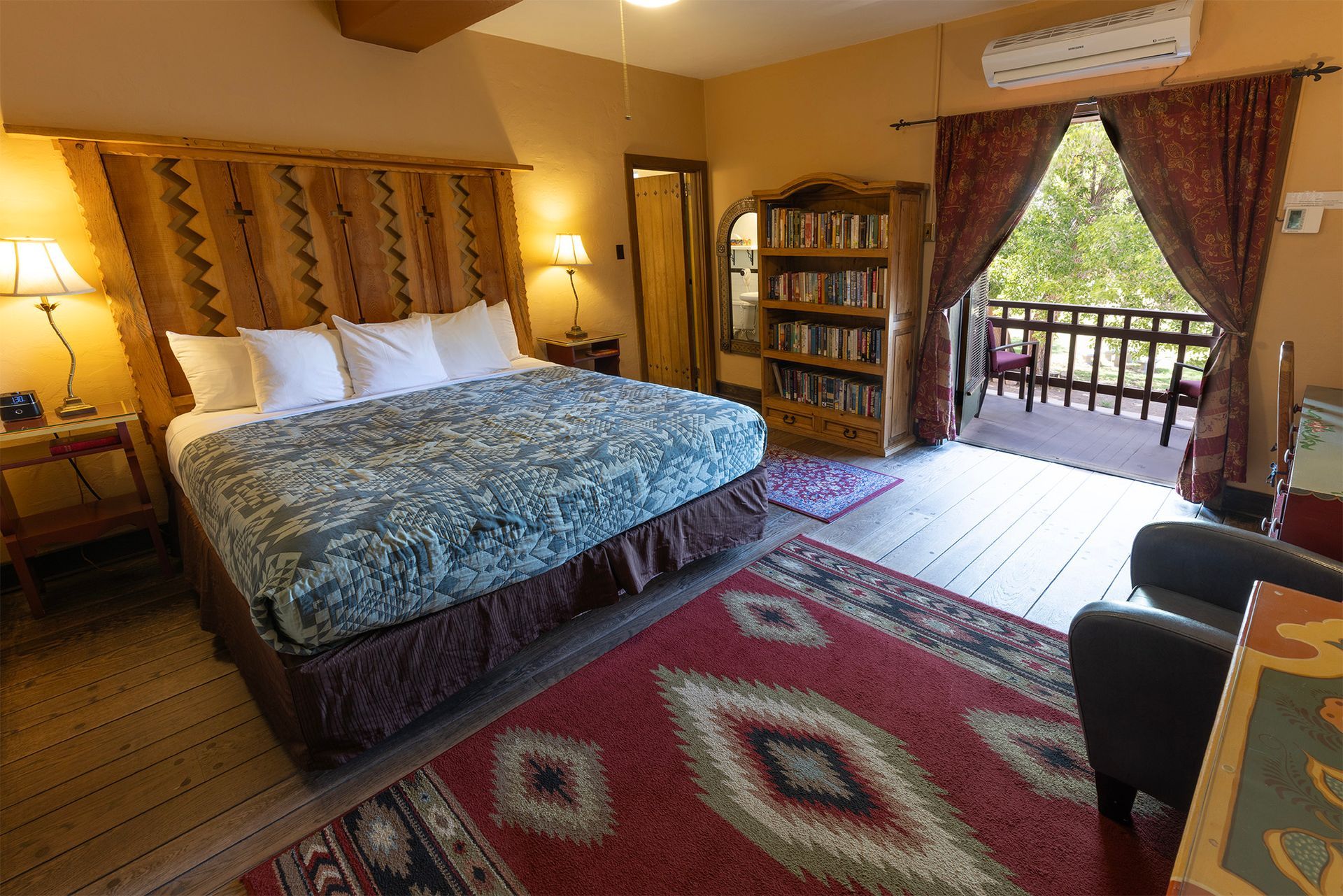 Bedroom with a king-sized bed, rug, bookcase, and balcony. Warm colors, natural light.