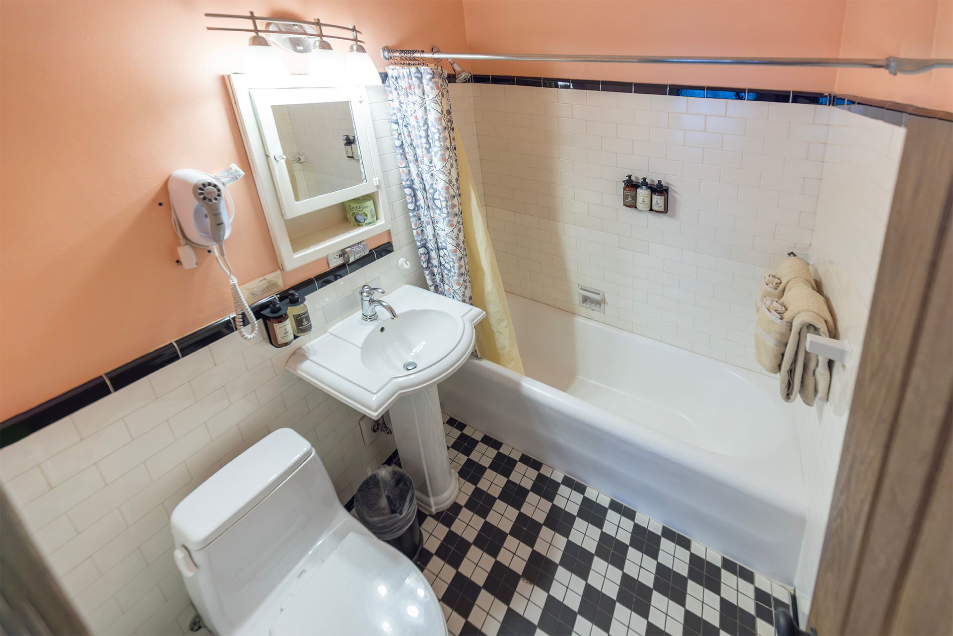 Small bathroom with a white tub and pedestal sink. Black and white checkered floor. Pink walls.