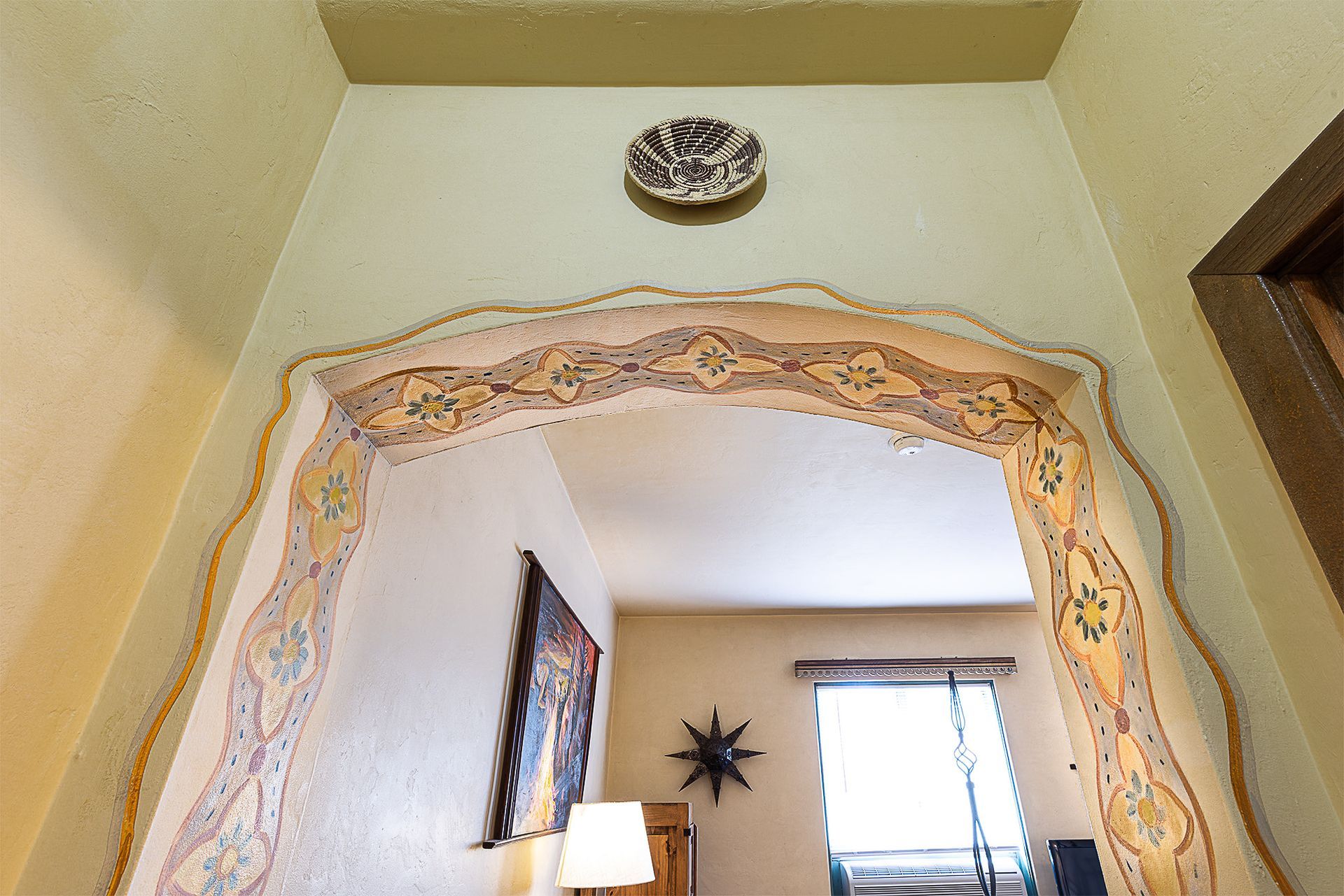 Ornate archway with detailed trim leads to a room with a painting, lamp, and window.